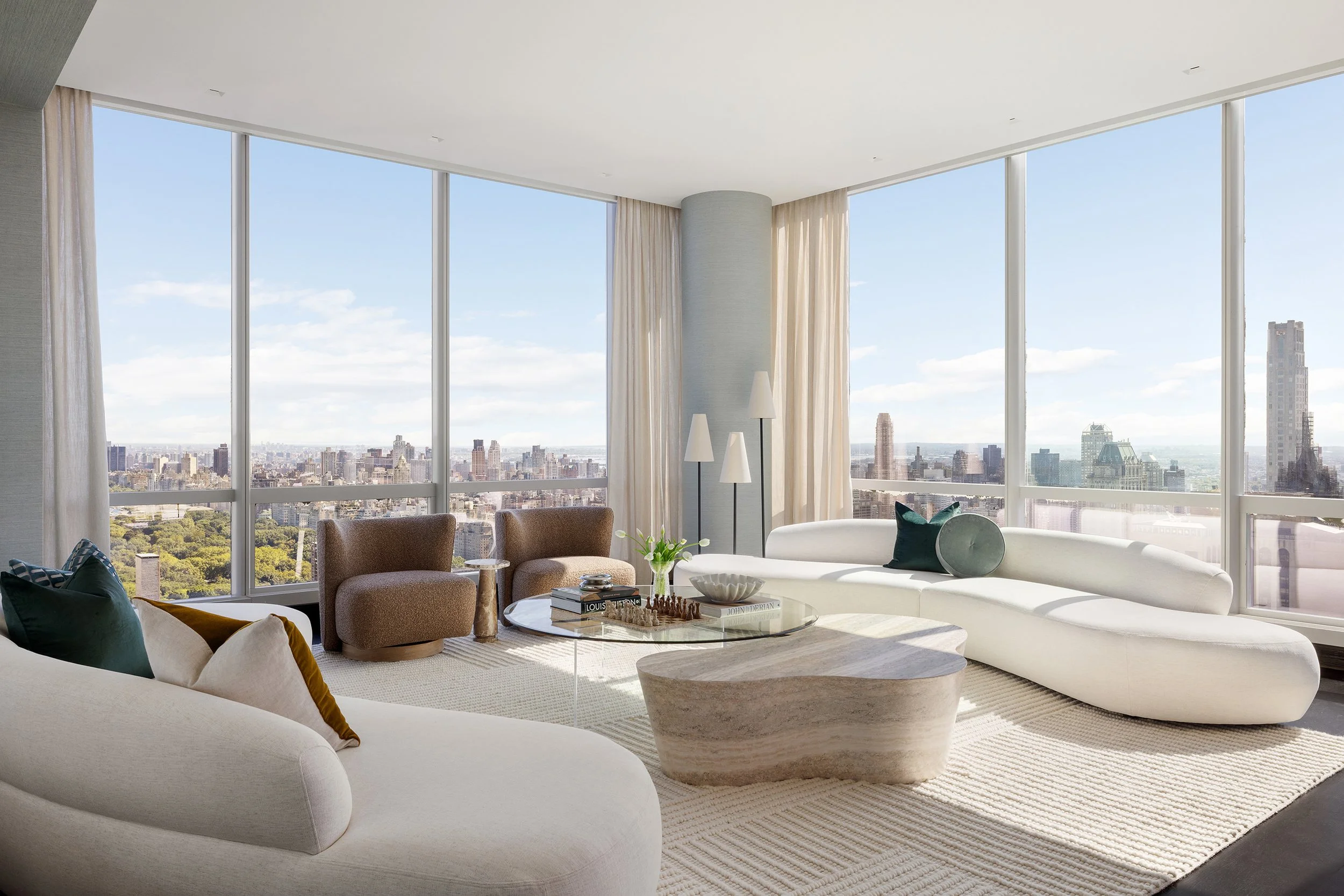 Luxury living room with large floor-to-ceiling windows, city skyline views, white curved sofa, beige armchairs, round coffee table, books, decorative objects, and modern floor lamps.