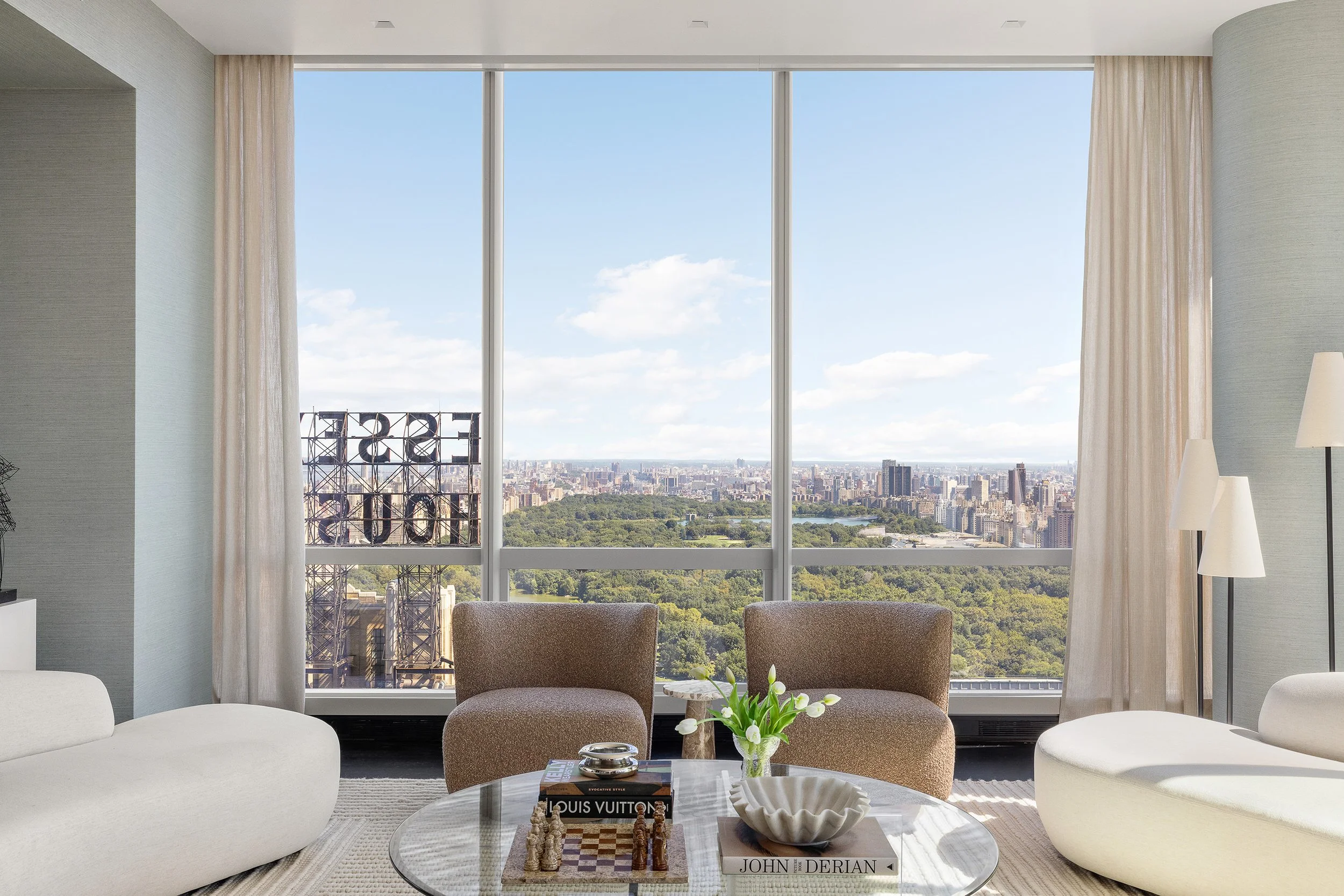 Living room with large window overlooking cityscape and central park, furnished with cream and beige chairs, a coffee table with books and decorative items, and a vase with white flowers.