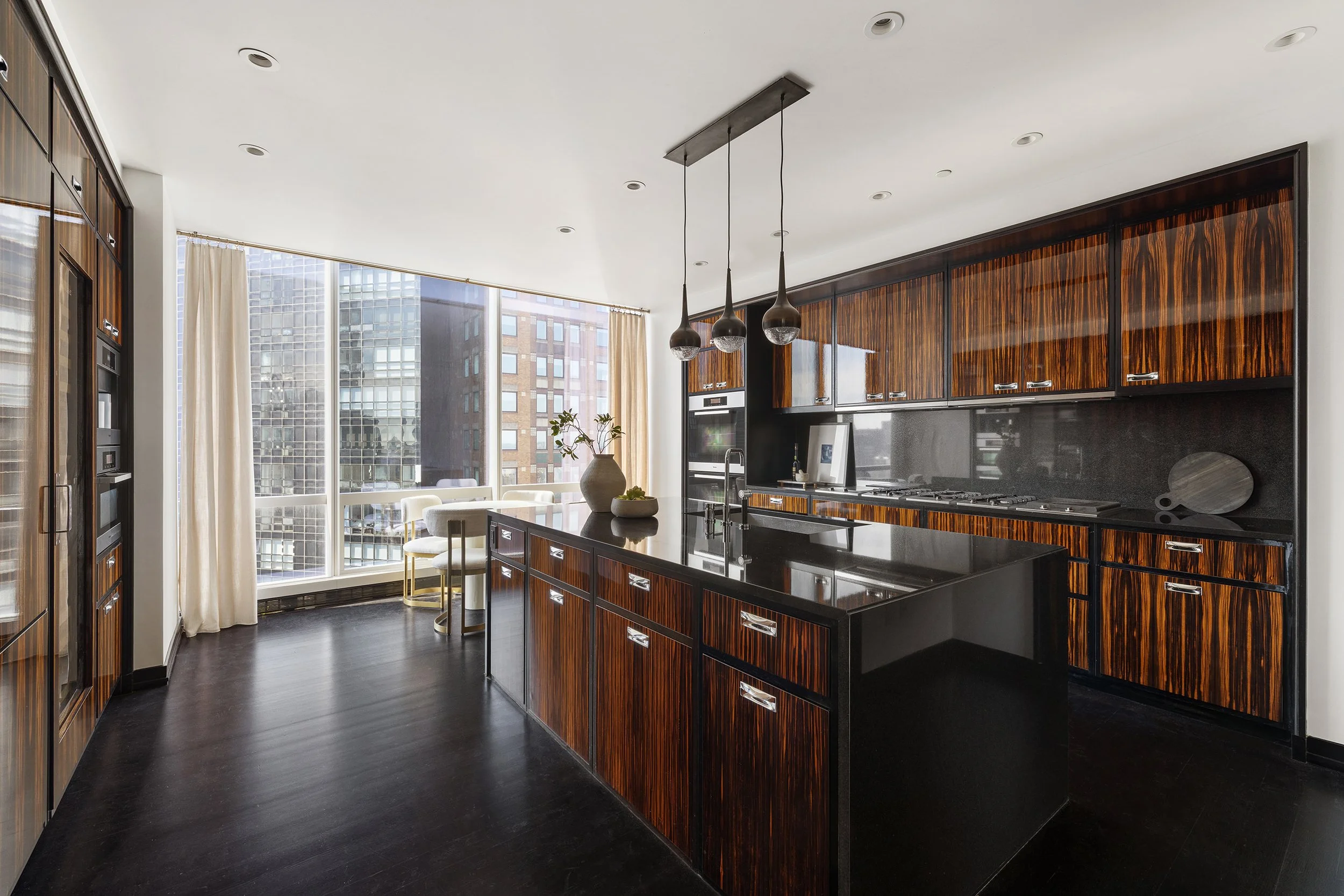 Modern kitchen featuring dark wood cabinets with a glossy finish, black countertop island, hanging pendant lights, and large windows with sheer curtains overlooking city buildings.