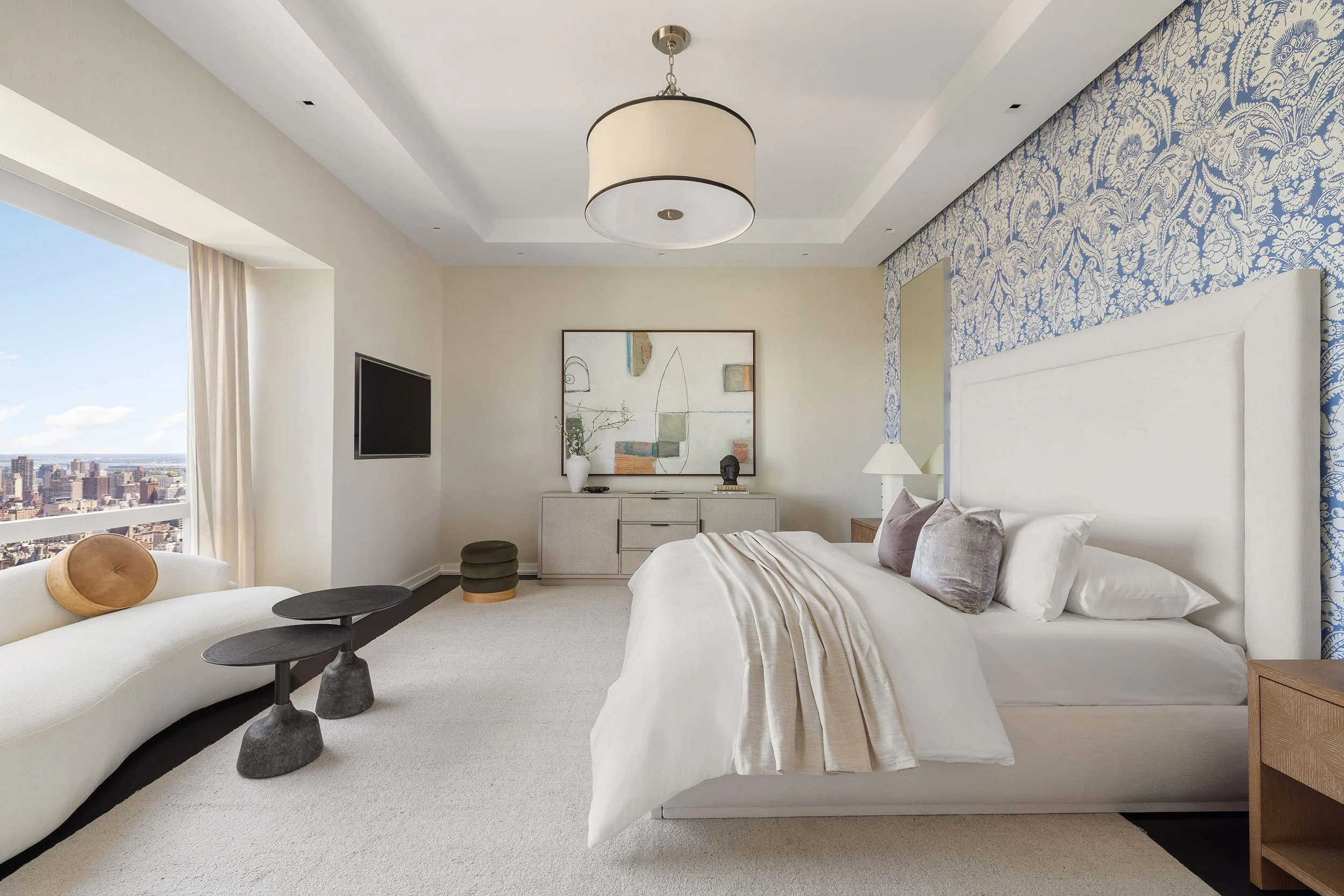 Modern bedroom with large bed, white headboard, gray and beige pillows, and beige blanket. There are two side tables with lamps, a white dresser with art and decor, a TV mounted on the wall, and a large window with city views and curtains.