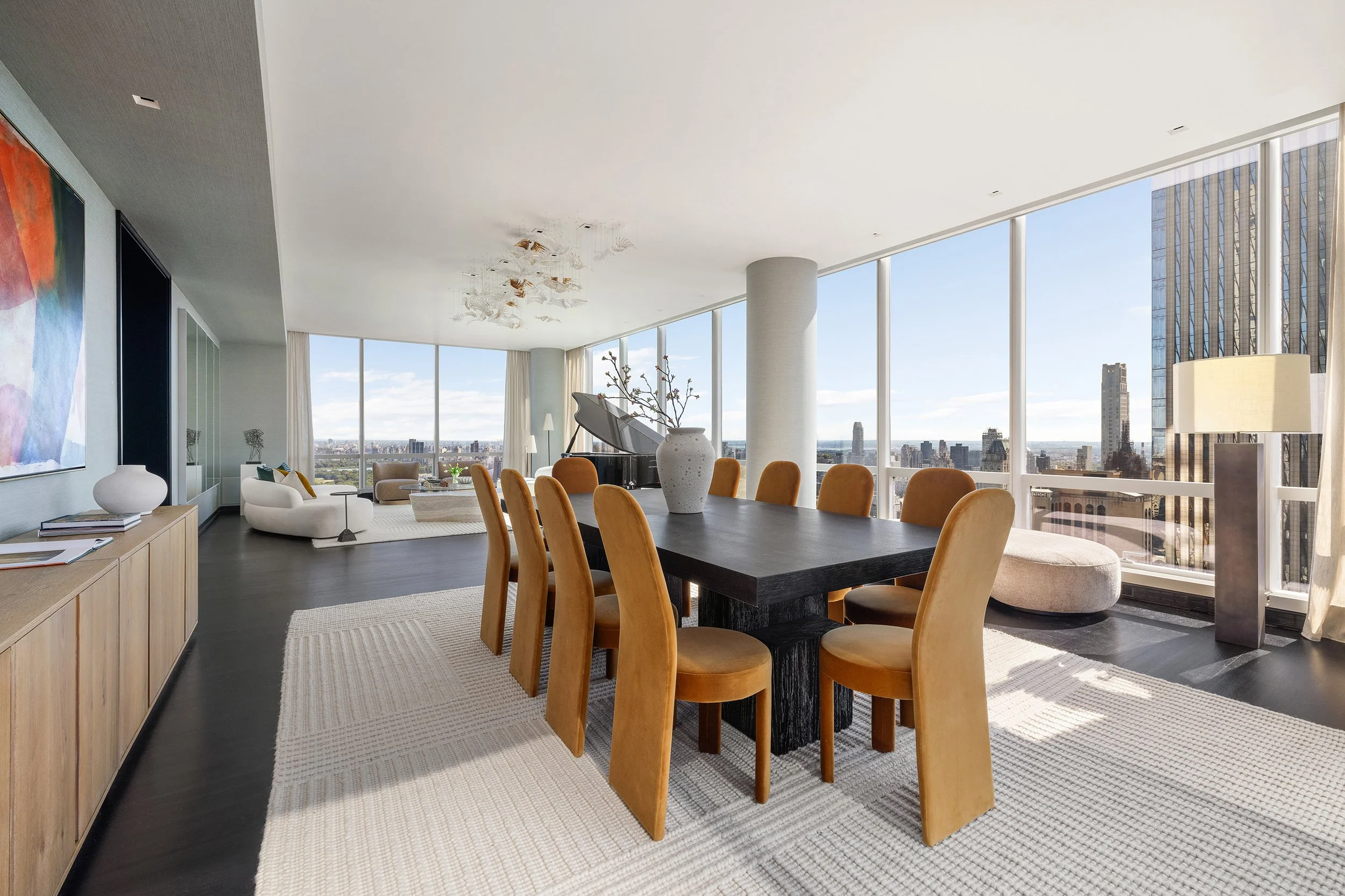 Modern high-rise apartment living room with large windows showing a city skyline, a round chill-out sofa, a grand piano, a large wooden dining table with yellow chairs, and colorful artwork on the wall.
