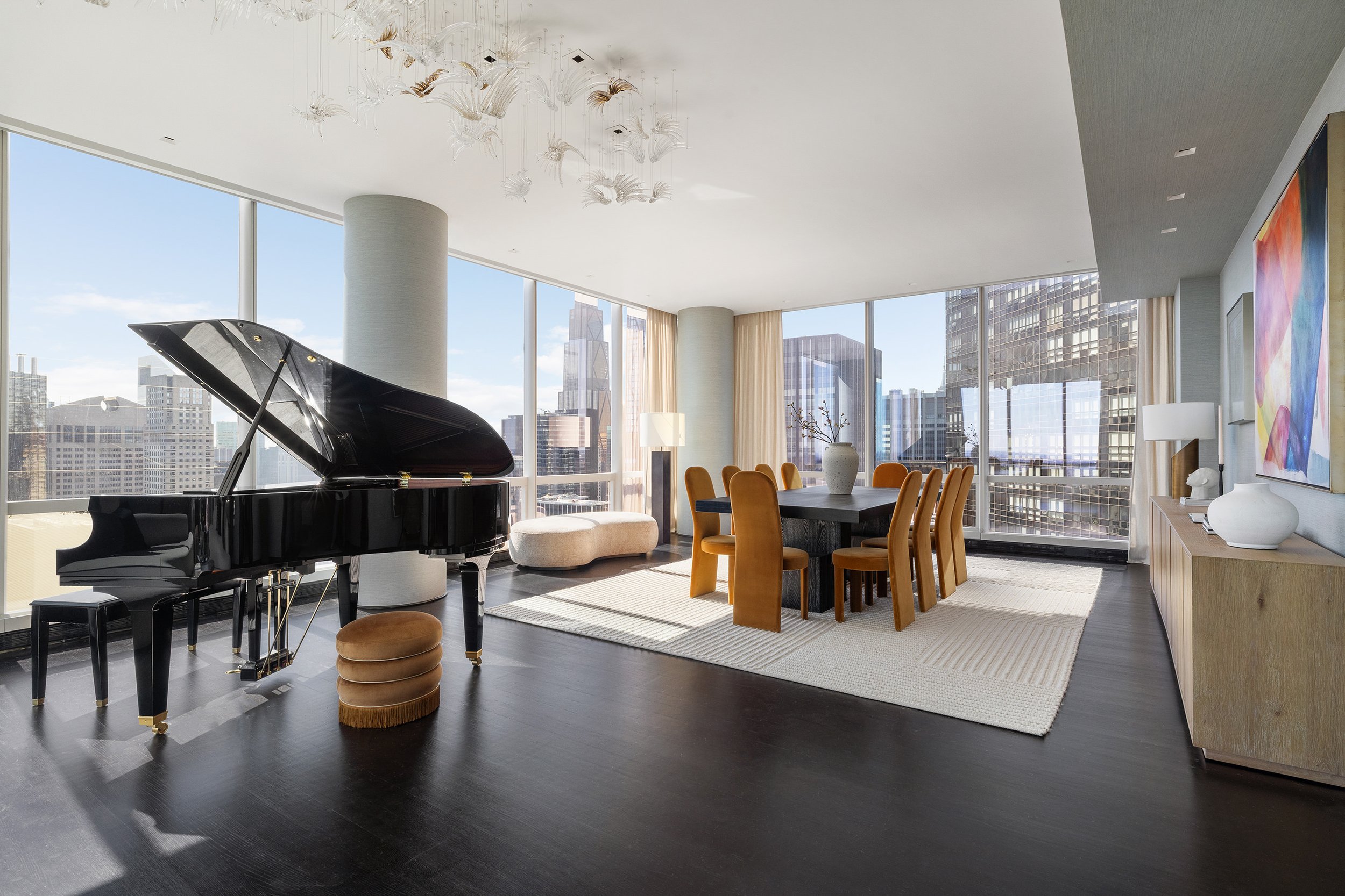 A modern high-rise living room with large floor-to-ceiling windows, a black grand piano, a white curved chaise lounge, a dining table with eight mustard-colored chairs, abstract artwork, and cityscape view.