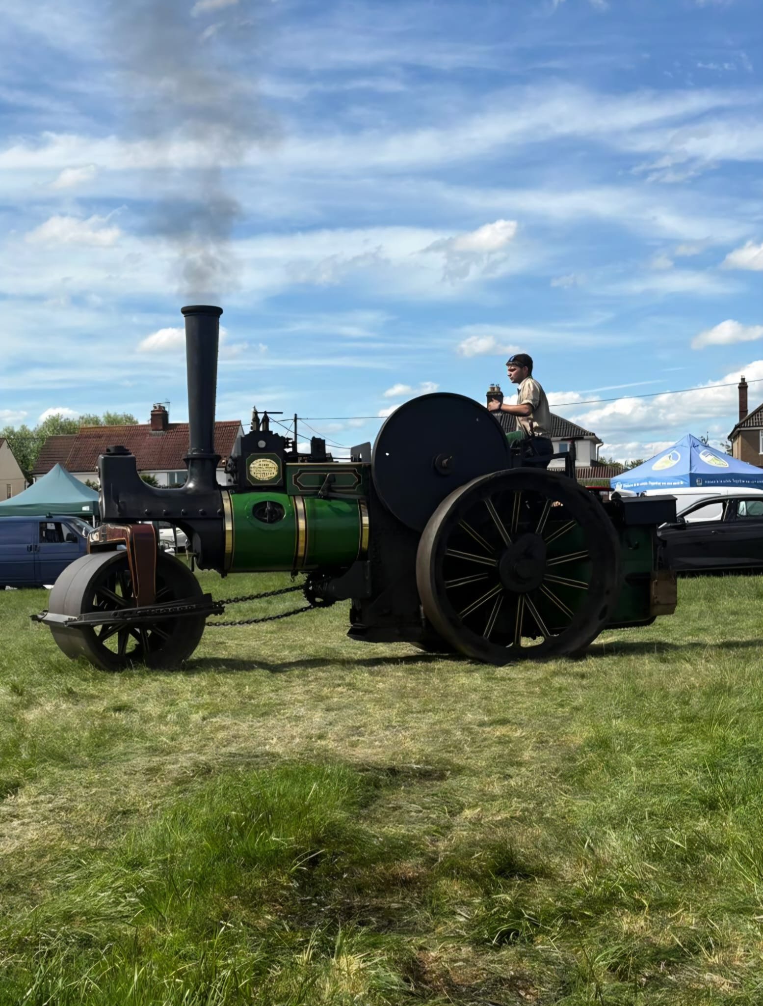 An outdoor scene with a vintage steam-powered tractor on a grassy field, under a partly cloudy blue sky, with rooftops and tents in the background.