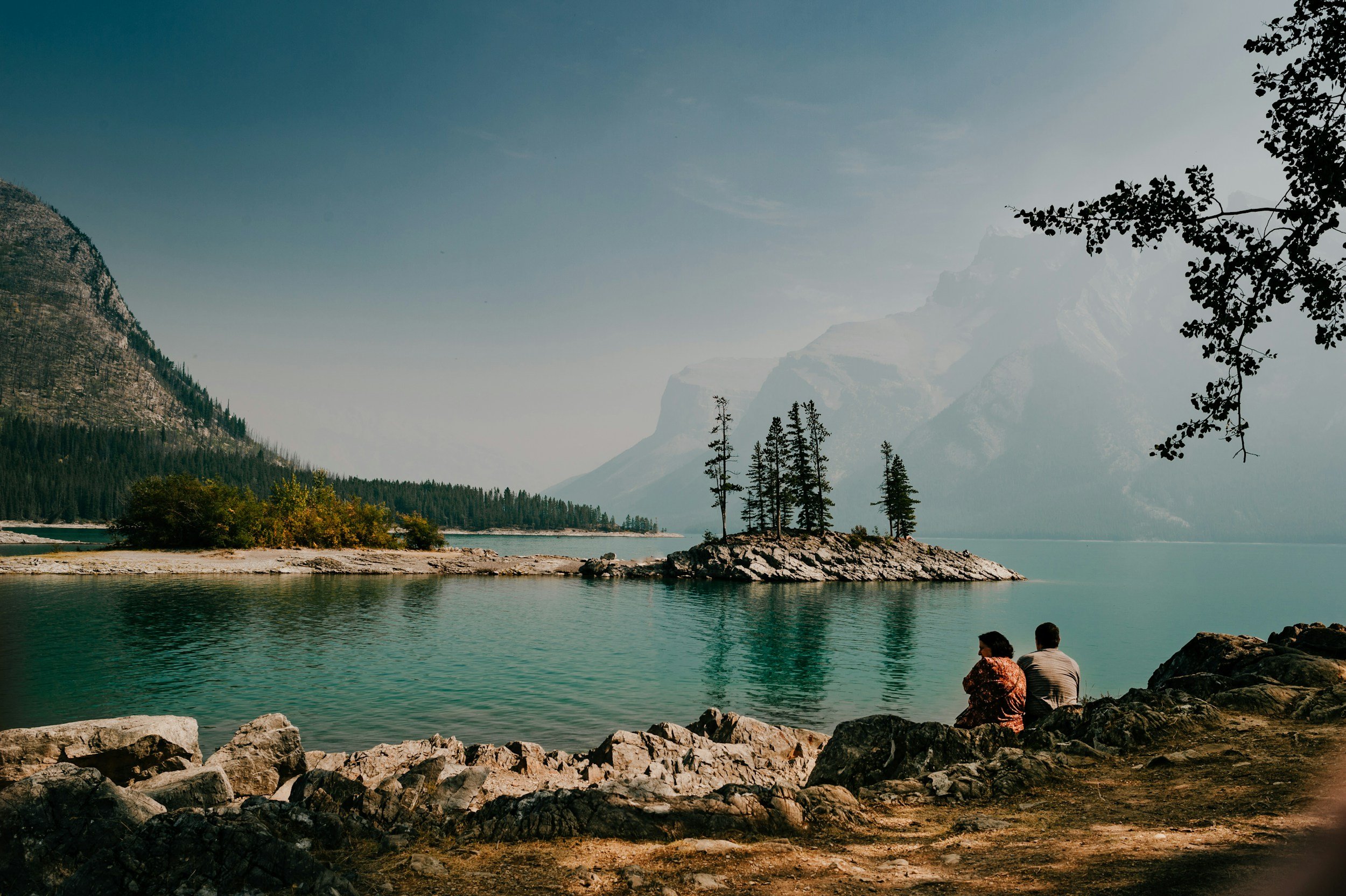 People sitting and relaxing by a lake surrounded by mountains and trees.
