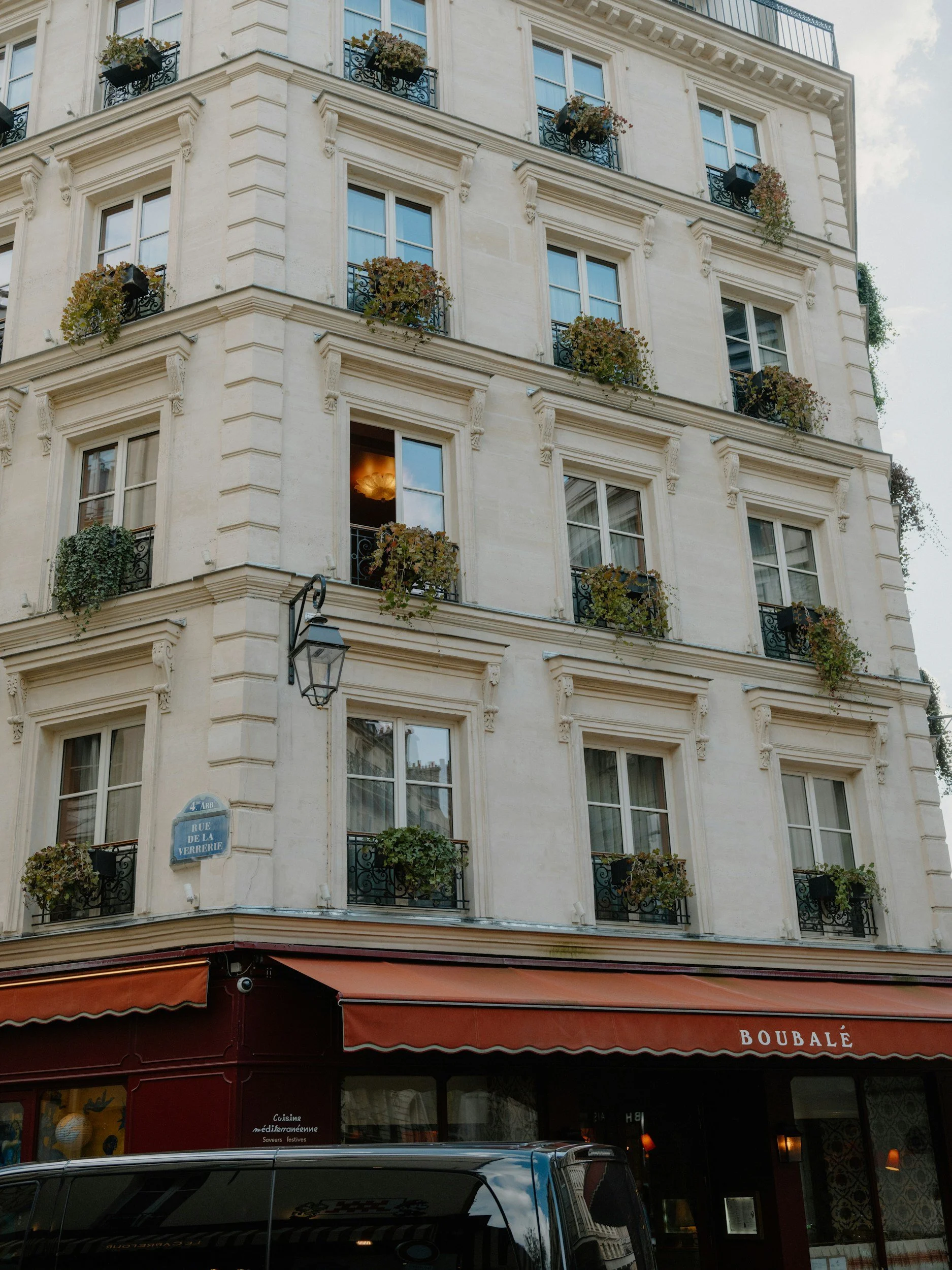 Corner of a Parisian building with multiple floors and large windows, decorated with potted plants and flowers. A street sign indicating 'Rue de la Verrerie' is visible, along with a storefront with a red awning labeled 'Boubalé.'