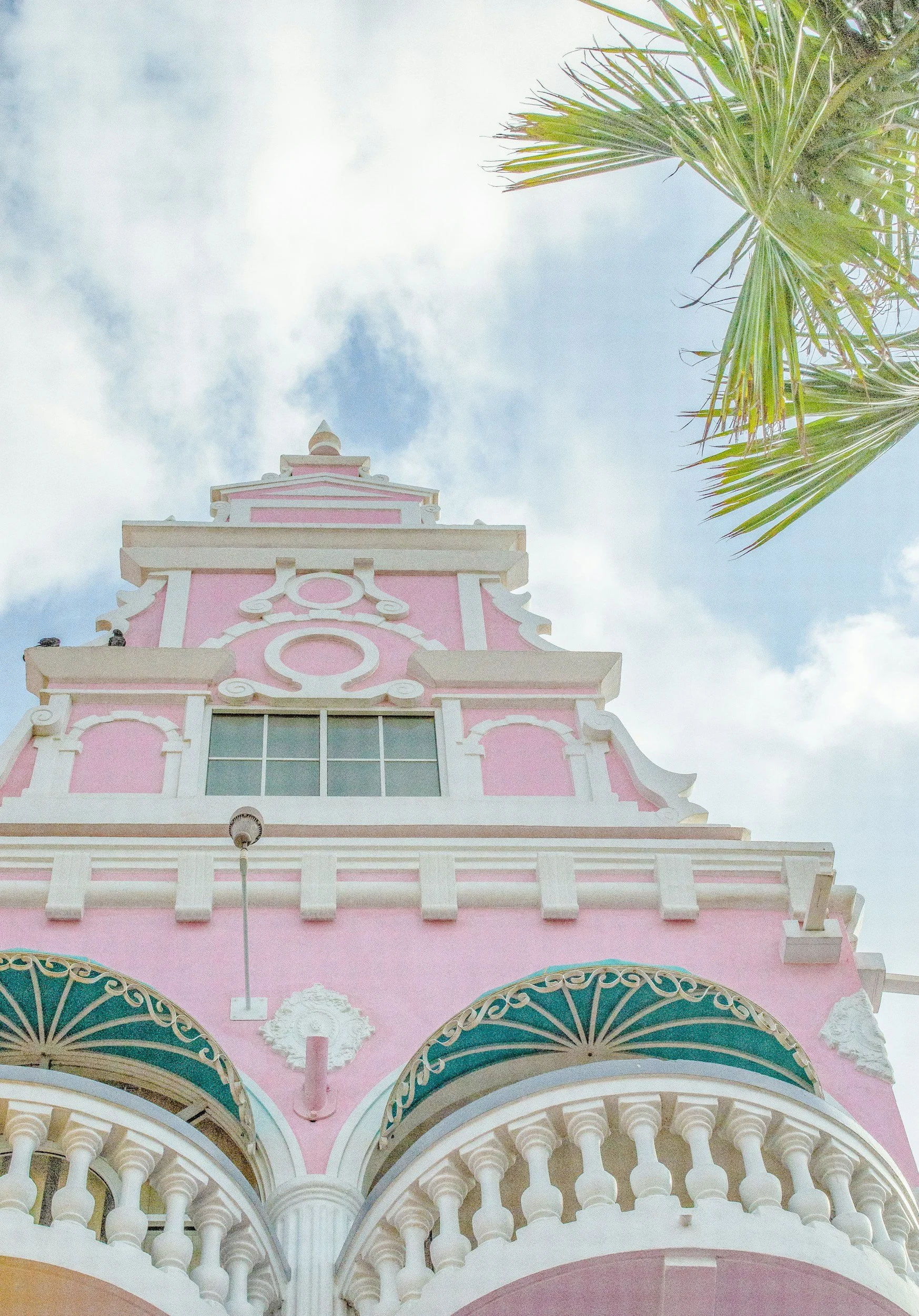 A pink and white building with ornate architectural details, including balconies, balustrades, and decorative moldings, with palm trees on the right side and a partly cloudy sky in the background.