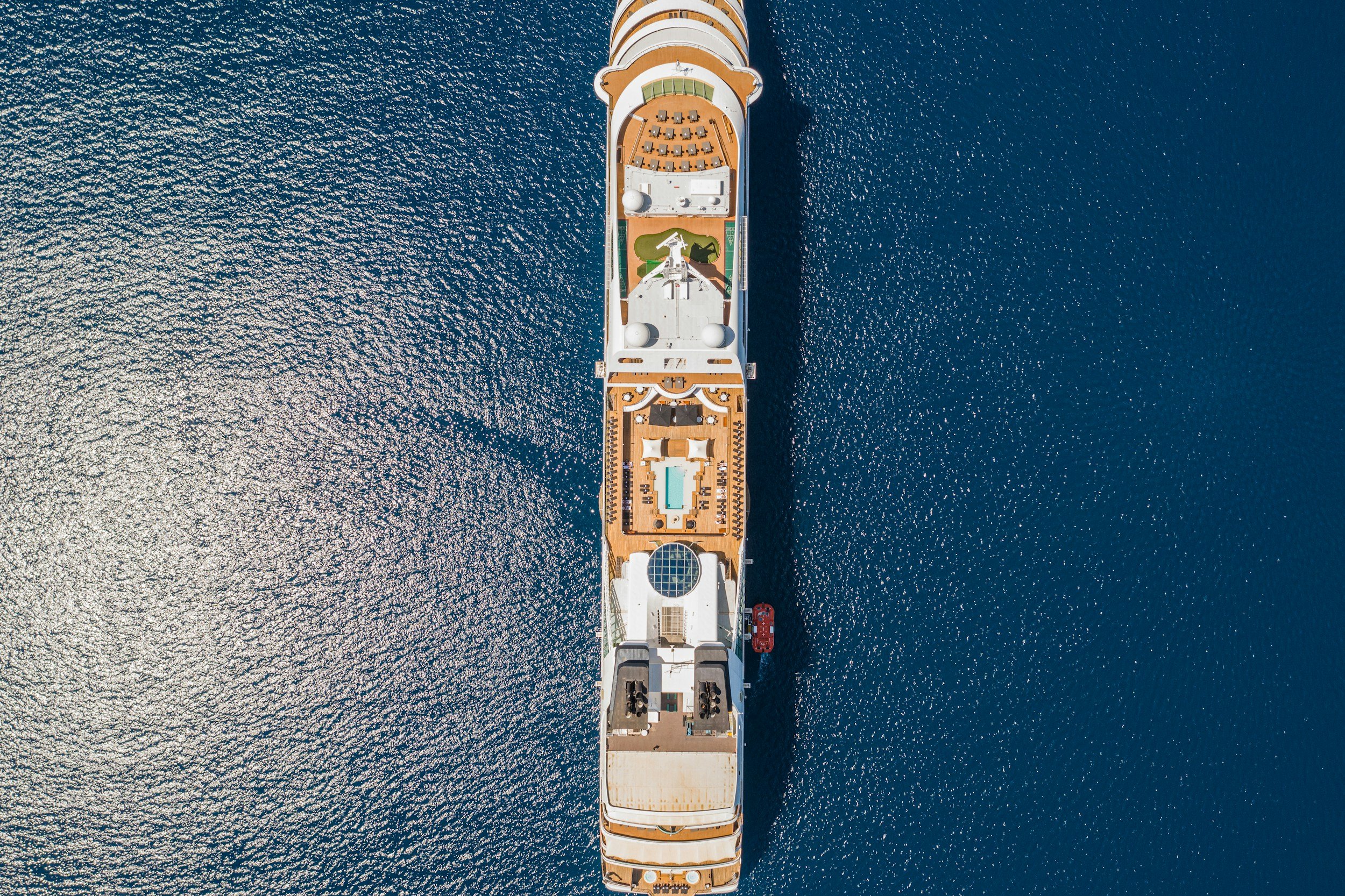 An aerial view of a cruise ship navigating in the open sea, showing the ship's top deck with lounge chairs, a swimming pool, and various amenities.