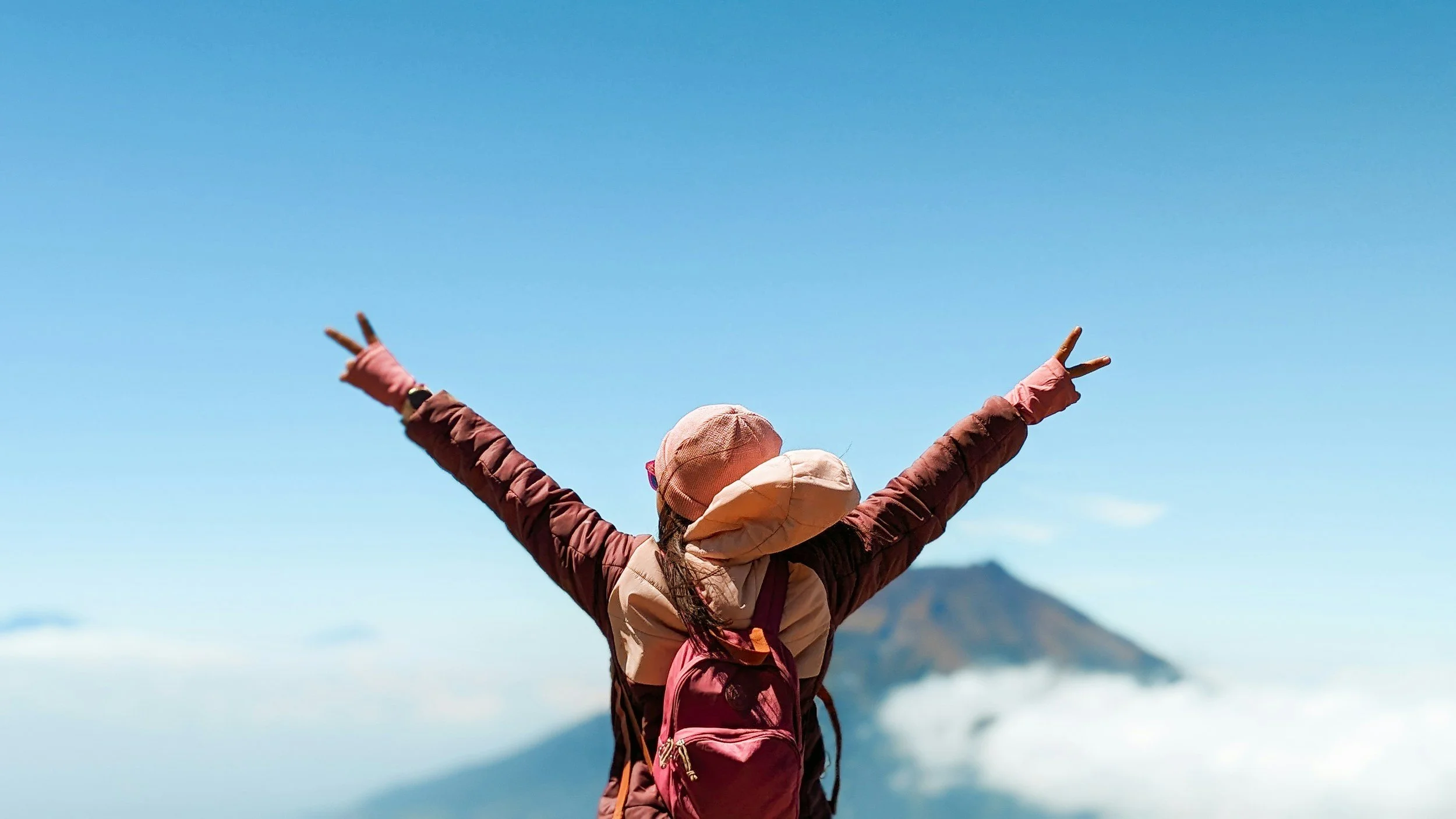 Person wearing a pink hat and pink jacket raising their arms in the air, facing a clear blue sky with mountains in the background.