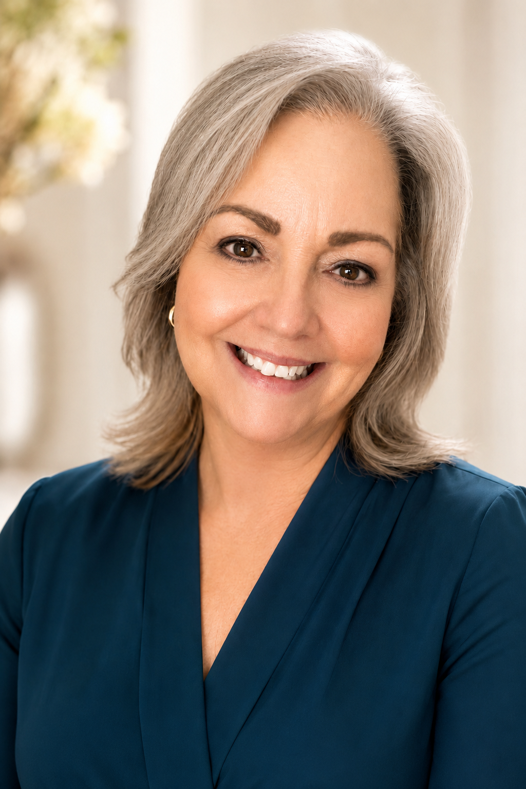 Smiling middle-aged woman with gray hair wearing a blue top, posed indoors with blurred background.