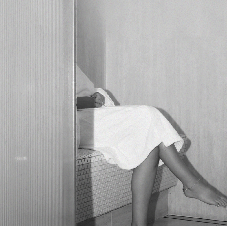A person sitting on a tiled bench with their legs hanging out, wearing a white dress and anklet, partially obscured by a wall.