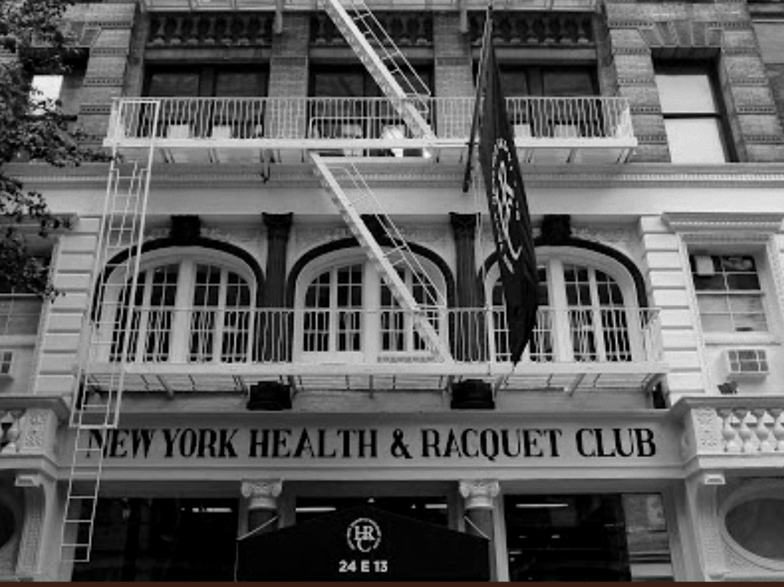 Black and white photo of a building with sign reading 'New York Health & Racquet Club'. Fire escape ladder on the left side of the building. Balcony with railings above the sign.