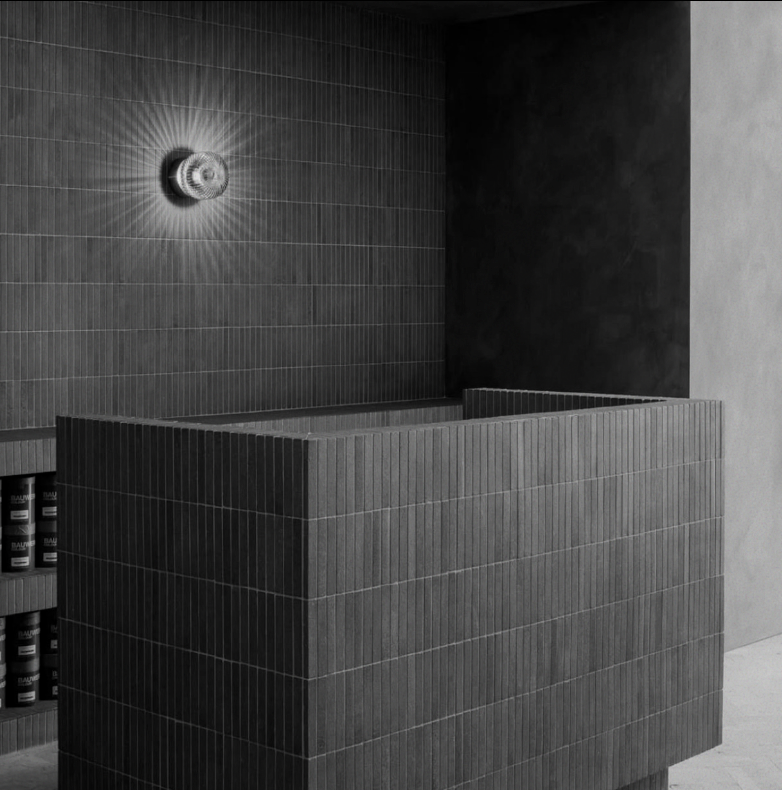 Modern minimalist bathroom with dark tiled walls, a dark tiled rectangular bathtub or sink, and a wall-mounted light fixture creating a starburst pattern.