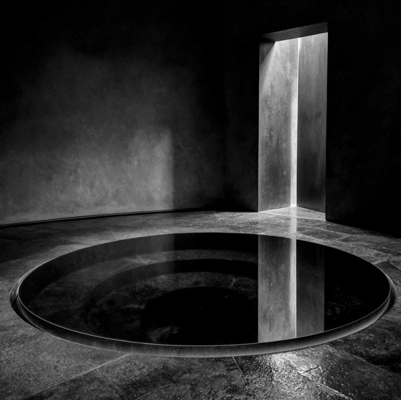 A black and white photograph of a minimalist interior with a large, circular, reflective black pool in the floor and light streaming in from a tall, narrow opening in the wall.