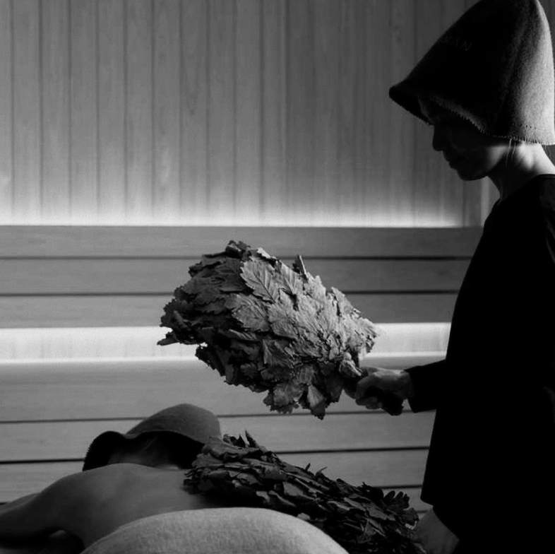 A person wearing a hoodie and glasses is holding a large bouquet of flowers, standing next to a seated individual in a relaxed posture, in a room with wood-paneled walls.