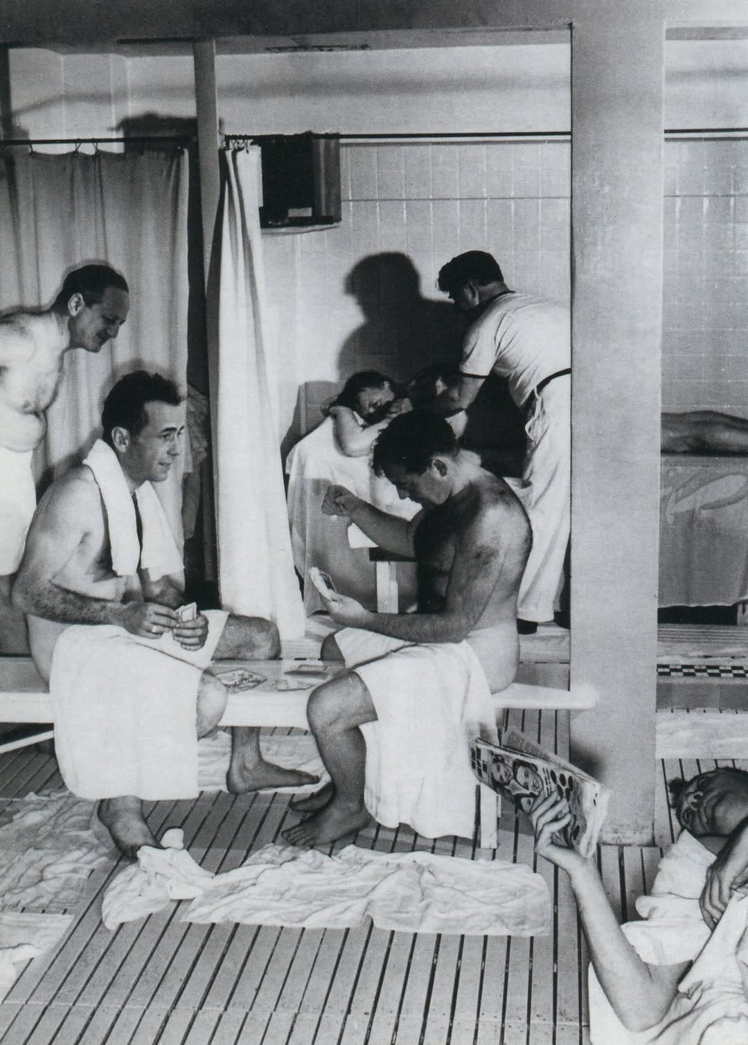 A group of people, including men and women, are in a hospital or medical care setting, with some sitting and others attending to patients, who are lying in beds and resting on the floor.