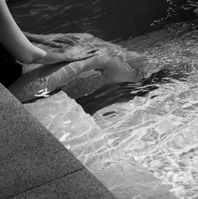 Two people holding hands in a pool, with one person's legs visible and the other person's hand resting on their knee.