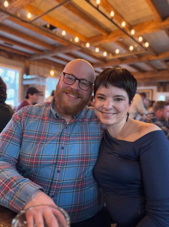 A smiling man with glasses and a beard, wearing a plaid shirt, sitting next to a smiling woman with short dark hair, wearing a dark blue top, in a warmly lit indoor setting with wooden beams and string lights.