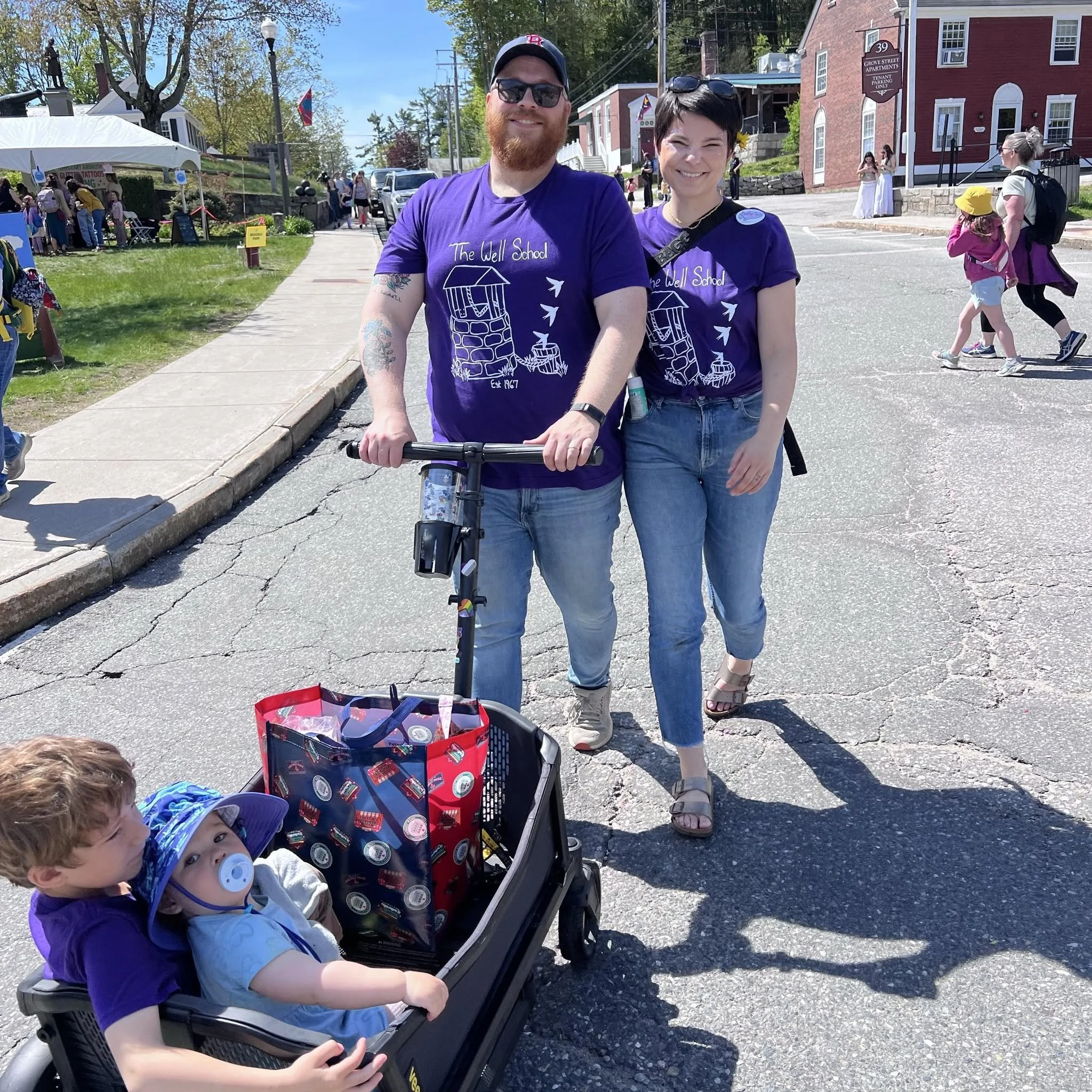 Two adults and two children at an outdoor event, with a woman smiling and a man with a beard wearing sunglasses and a purple T-shirt, standing next to a stroller with a child sitting inside. The child in the stroller has curly hair, a pacifier, and a blue hat. The background shows other people, tents, and buildings on a sunny day with trees and a blue sky.