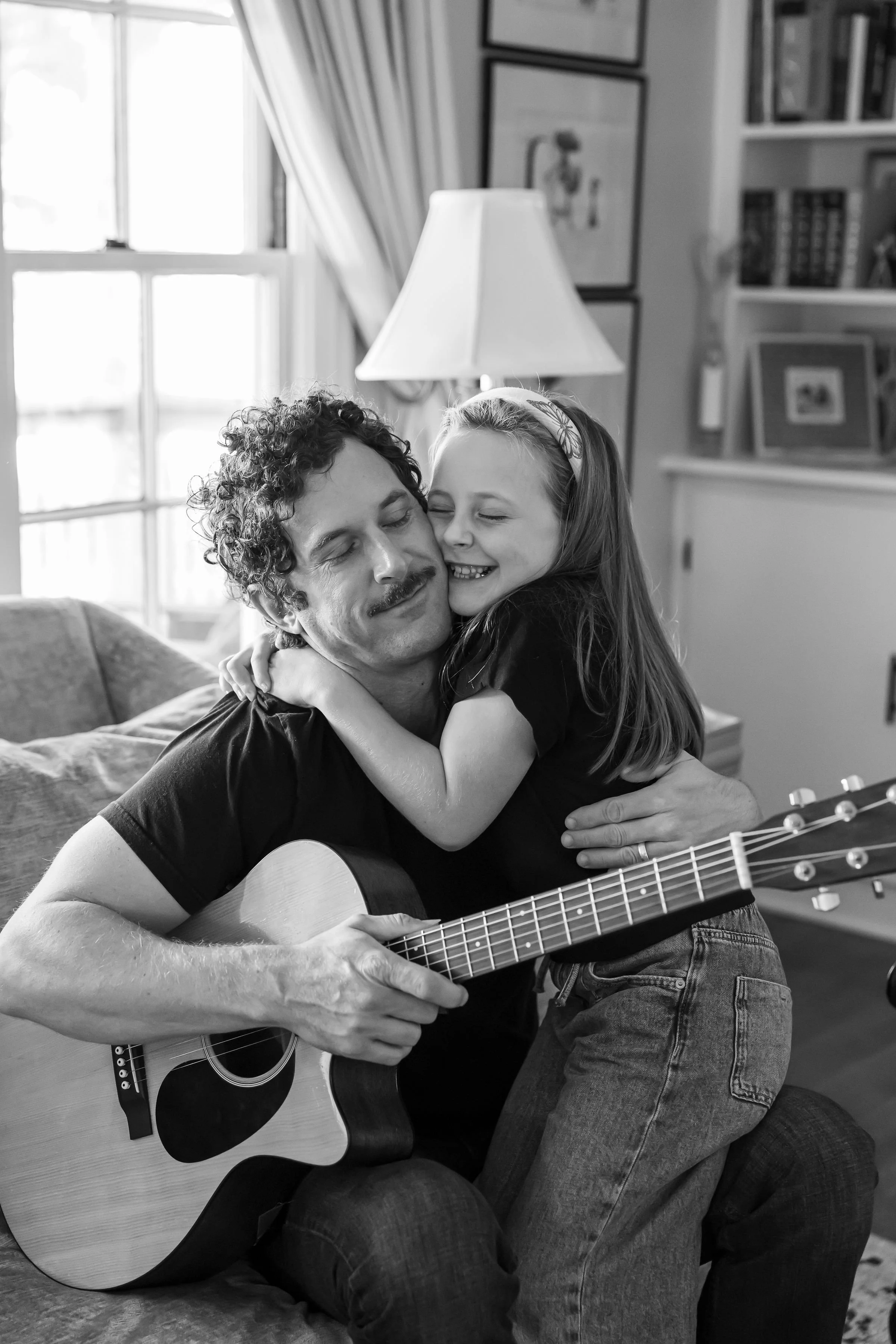 A man playing an acoustic guitar and a young girl hugging him, both smiling, inside a cozy room with a window, bookshelf, and artwork in the background.