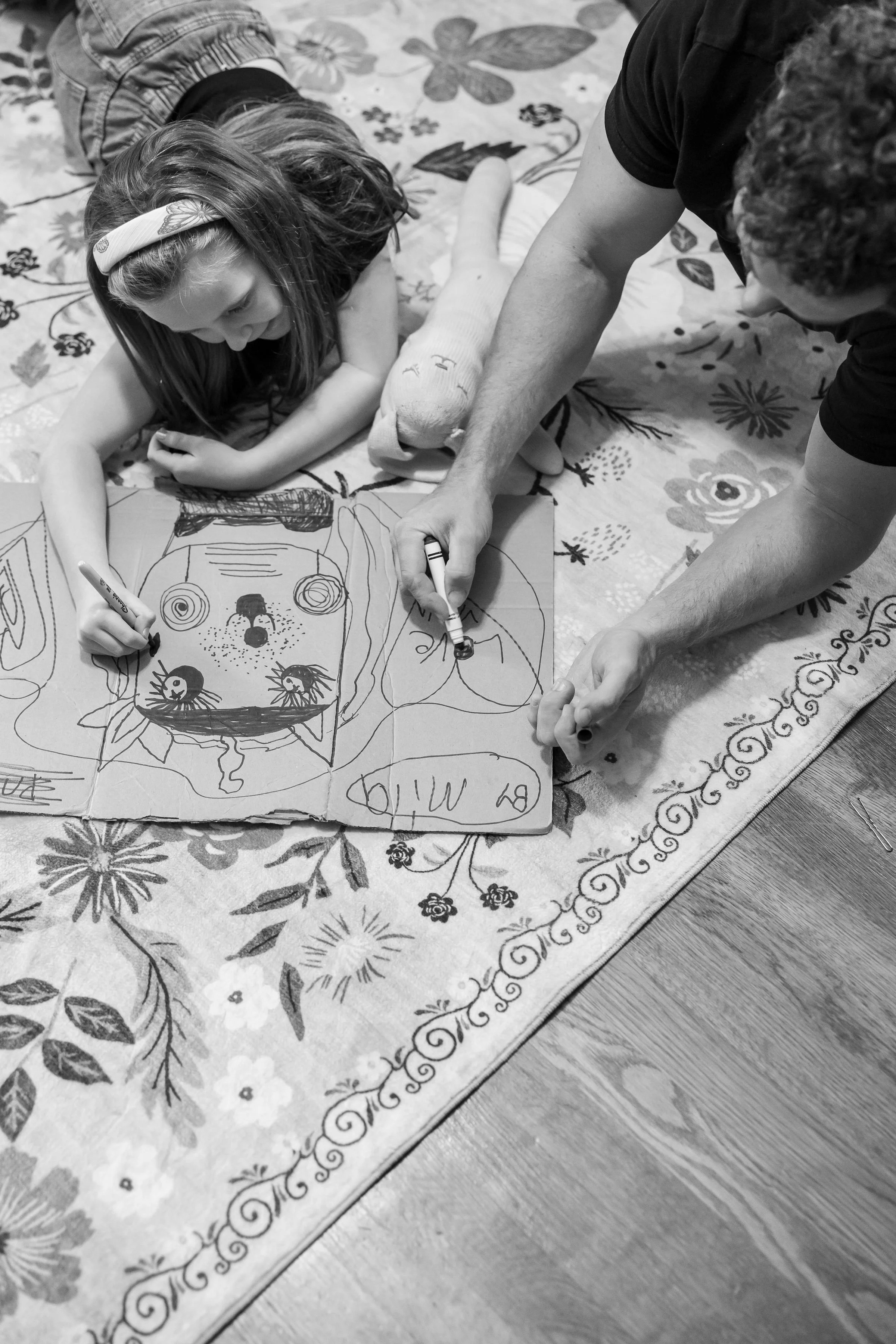 Child and adult drawing and coloring a large poster of a dog on the floor, on a floral rug.