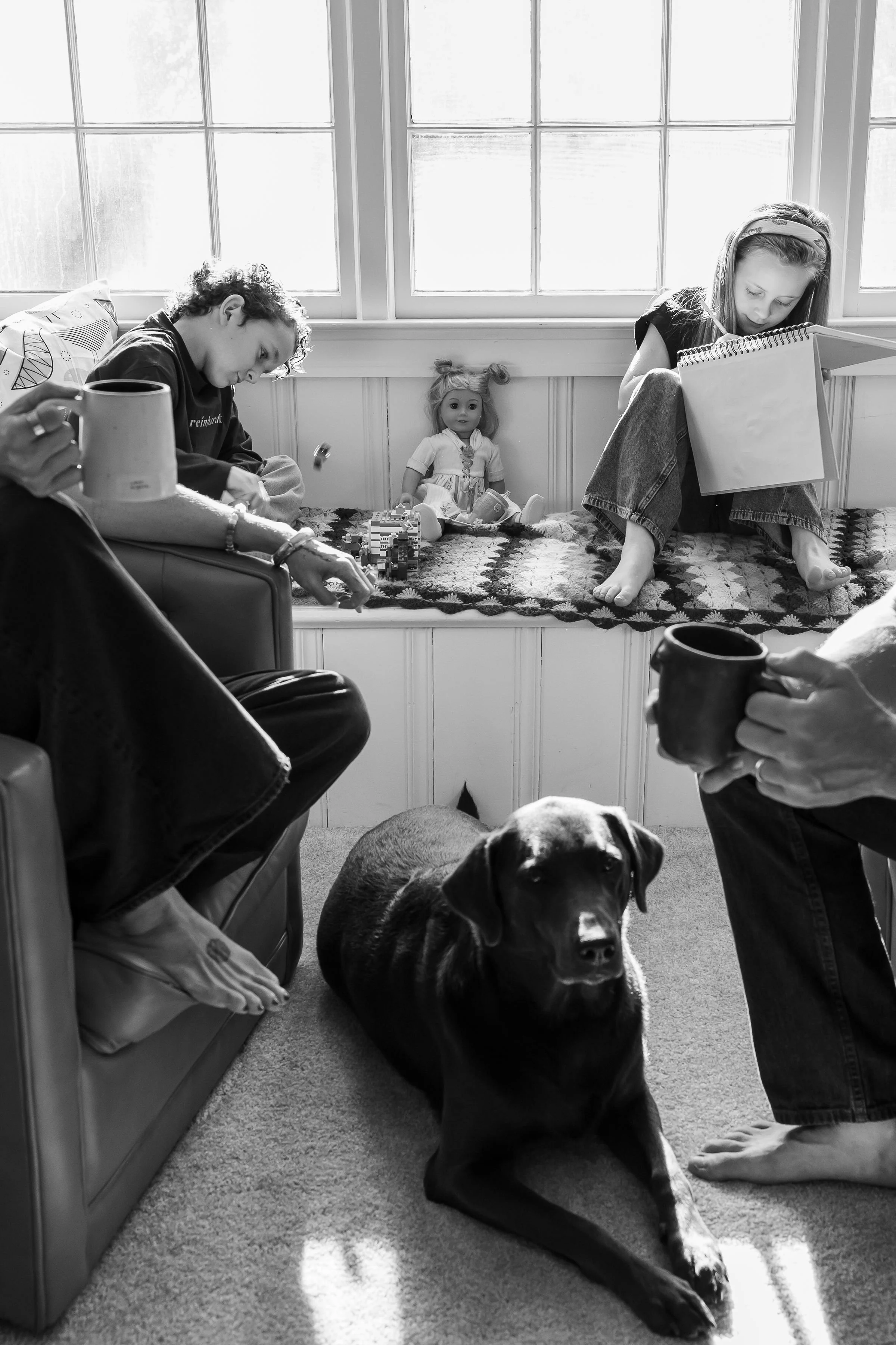 A young boy and a young girl sit on a window seat in a sunlit room. The boy is playing with small toys or building blocks, and a doll is positioned between them on the seat. The girl is engrossed in drawing or reading a large notebook. A large dog, possibly a Great Dane, is lying on the carpet in front of them, looking directly at the camera. The scene is cozy, with natural light streaming through large windows, and the room has a carpeted floor and a patterned blanket on the seat.