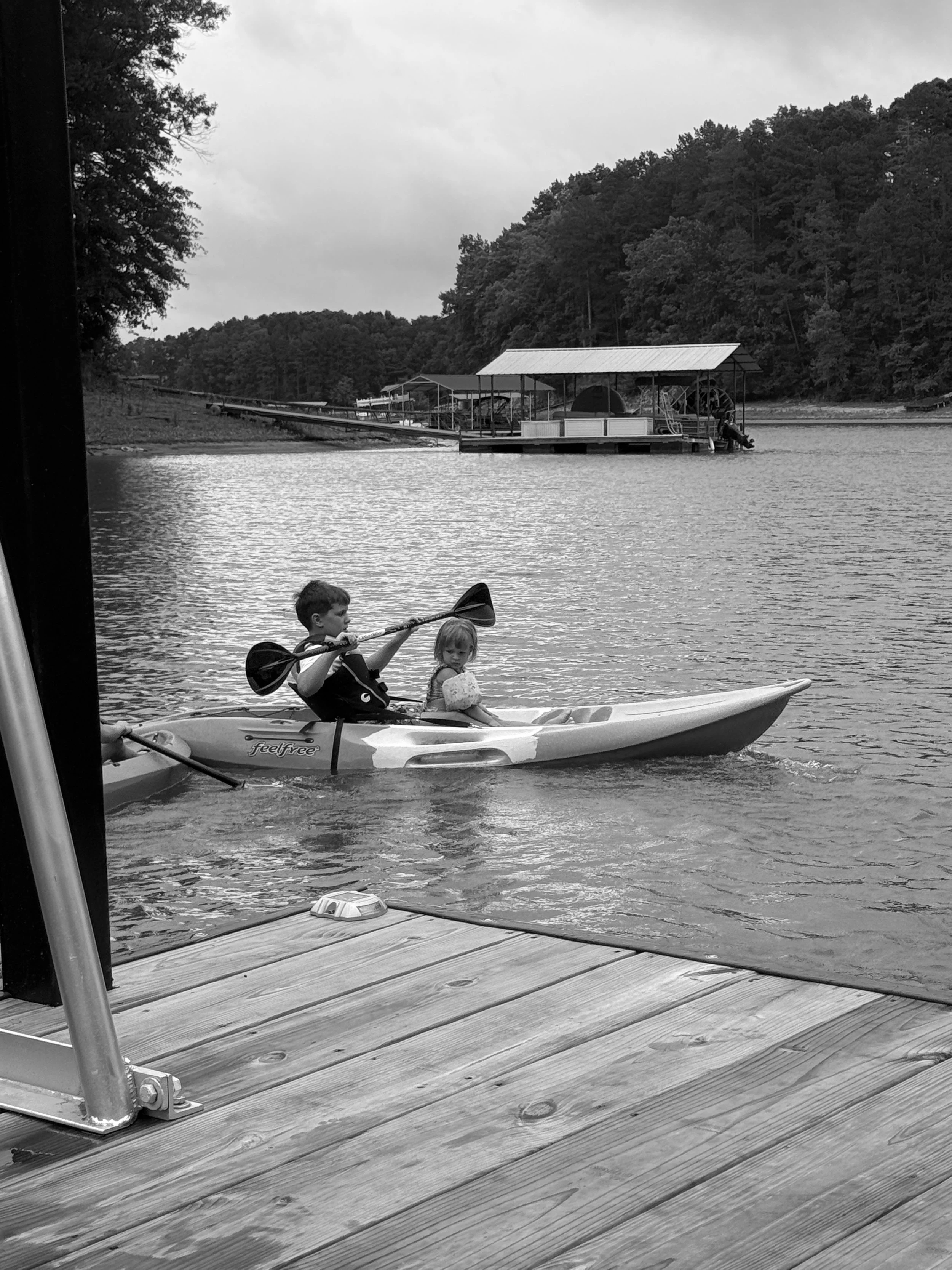 Children kayaking on a lake near a wooden dock with a floating boat house and a forested hill in the background.