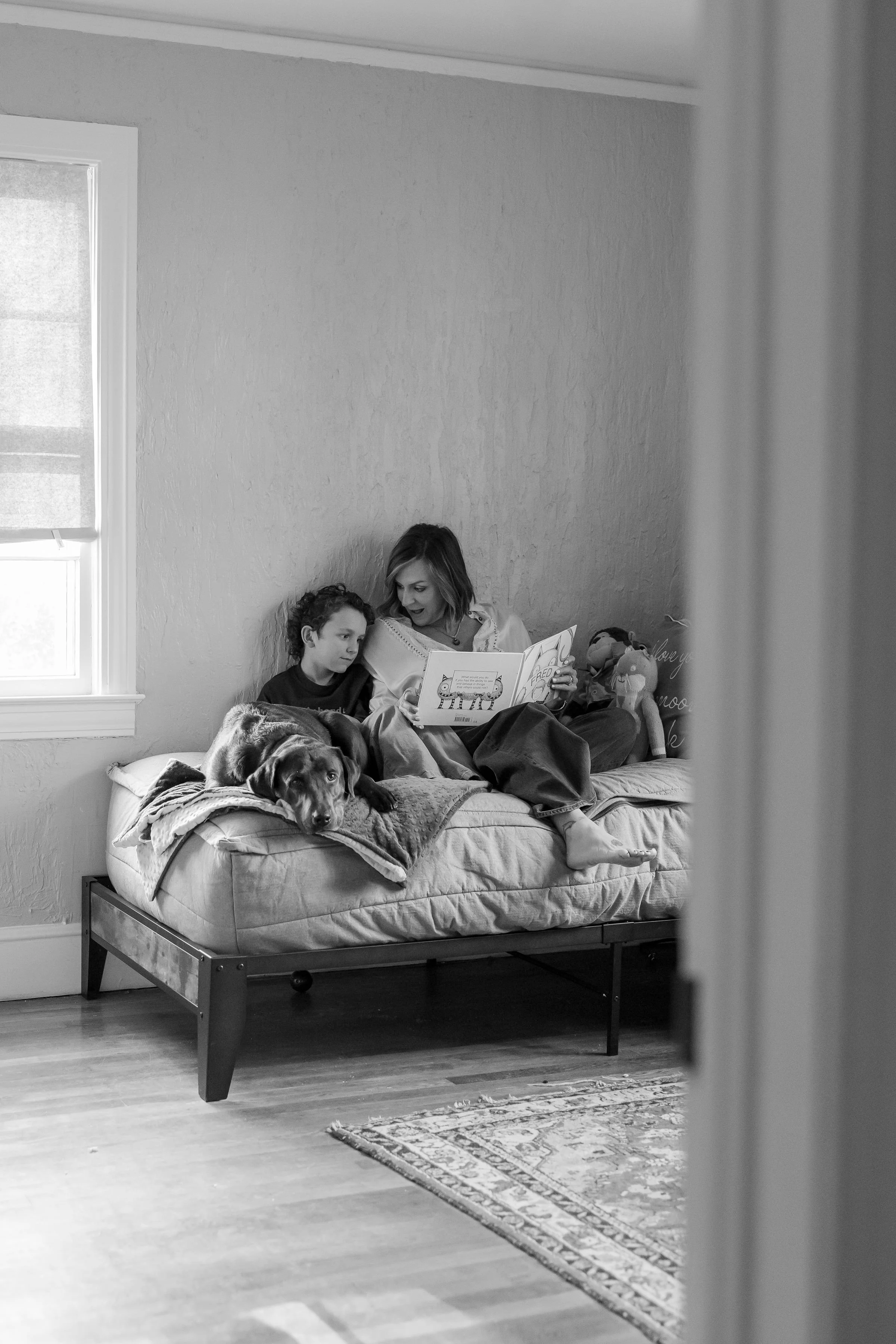 A woman and a boy sitting on a bed, reading a book together, with a large dog lying beside them on the bed. The room has a window with closed blinds, a patterned rug, and a decorative pillow with writing on it.