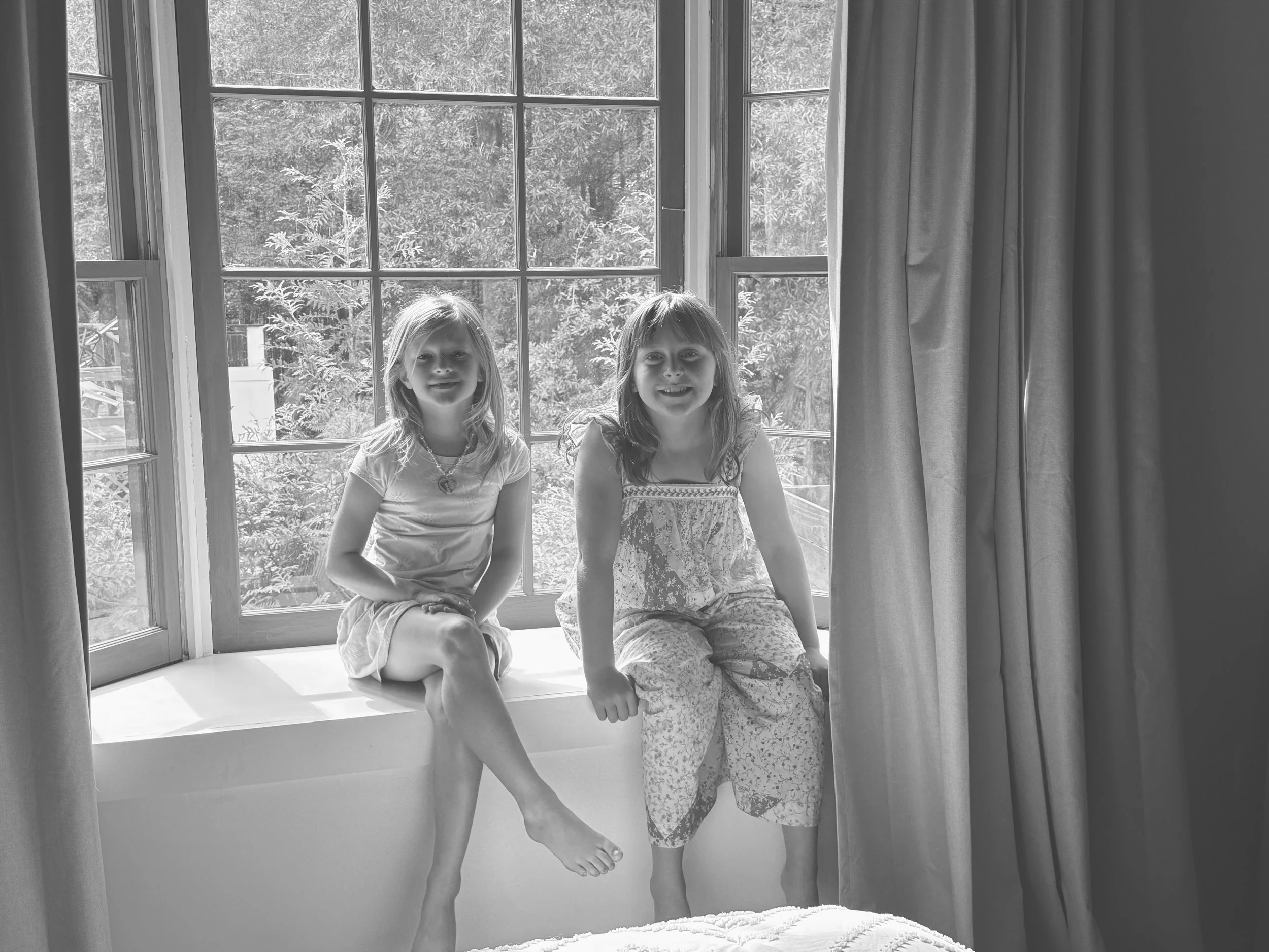 Two young girls sitting on a window seat by a large window with curtains, with trees visible outside.