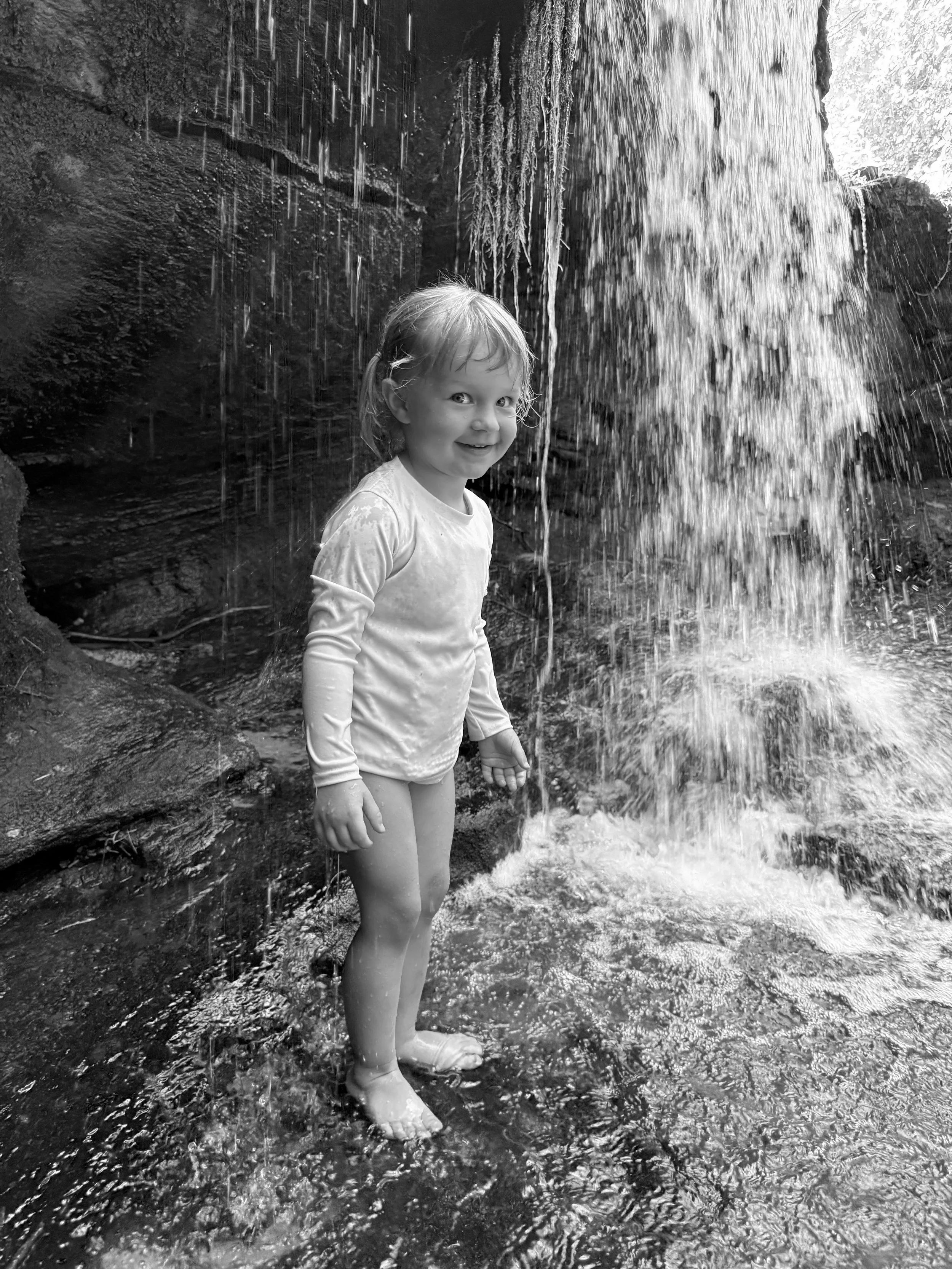 A young girl standing in a shallow stream under a waterfall, smiling and looking at the camera. She is wearing a long-sleeved shirt and rain boots, with her wet hair sticking to her face. The background shows the rocky sides of the creek.