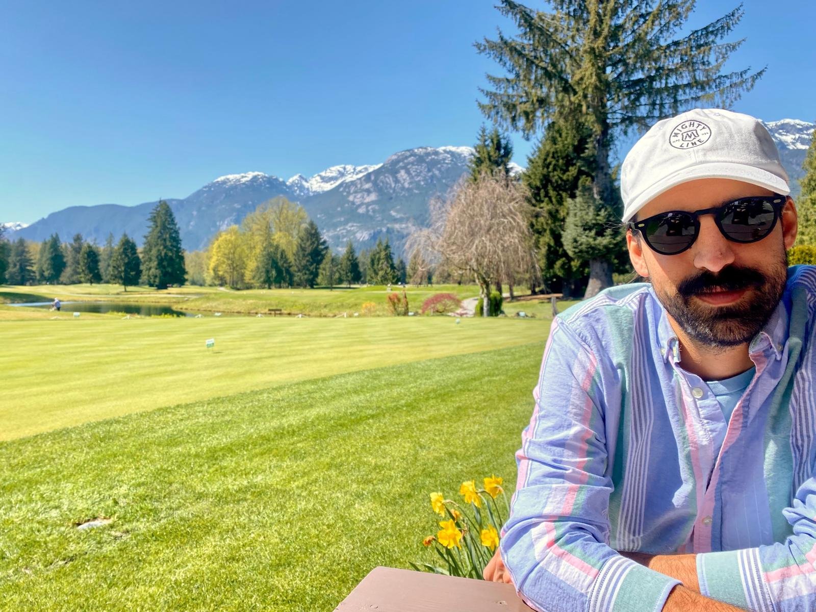 Daniel Deal wearing sunglasses and a white cap sitting outdoors on a bright, sunny day with a mountain range and trees in the background.