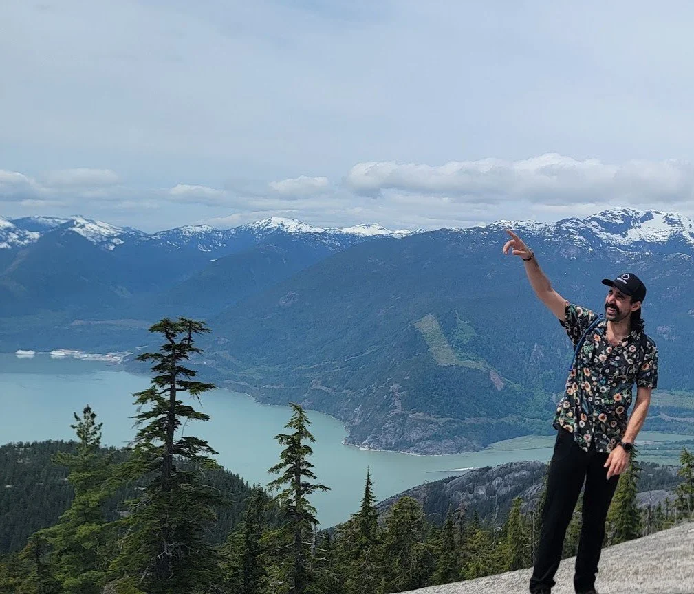 Daniel Deal with a wide smile, wearing a floral shirt and black cap, stands on a mountain overlook pointing towards snow-capped mountains and a large turquoise lake surrounded by forested hills under a partly cloudy sky.