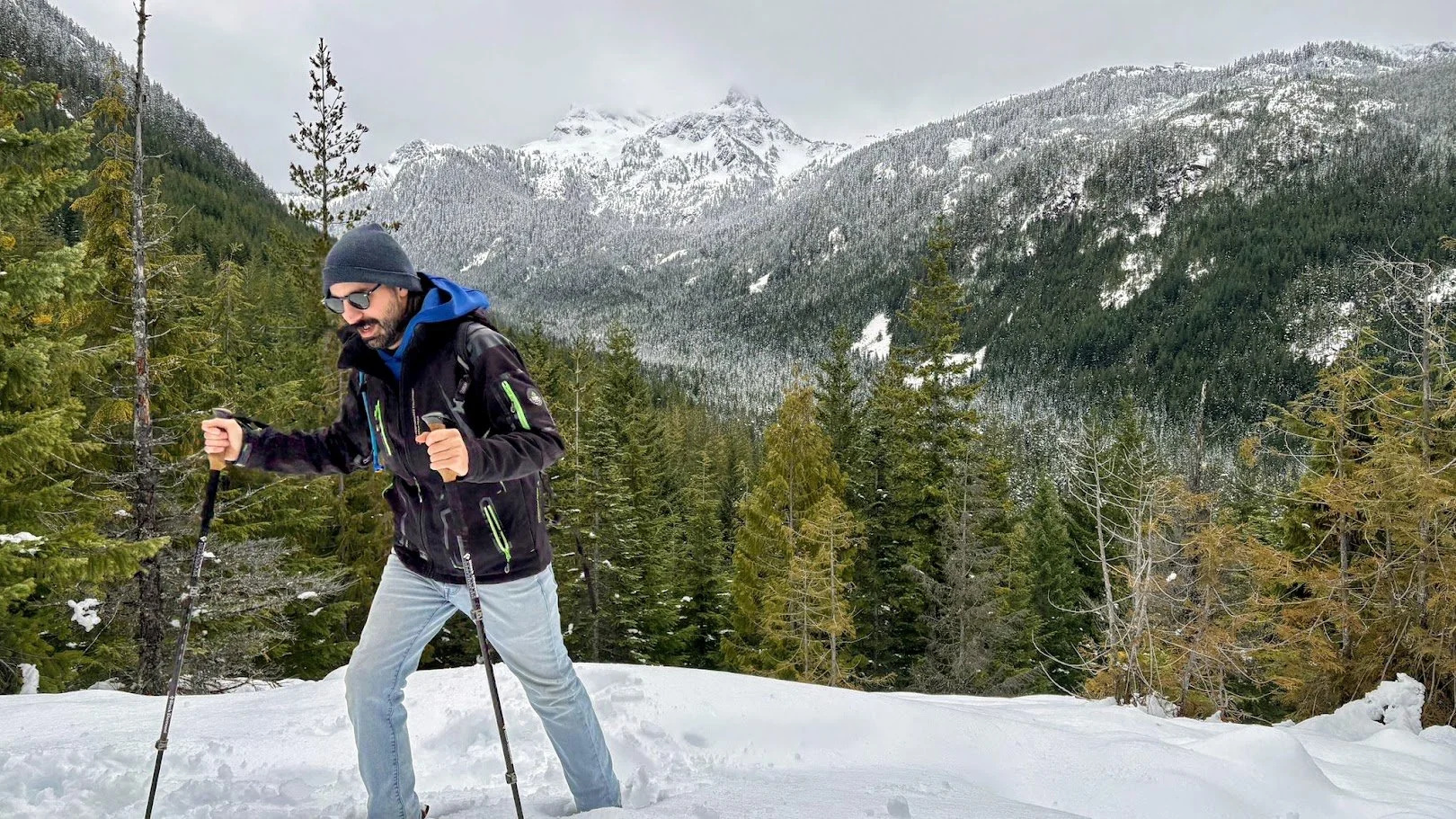 Daniel Deal hiking with trekking poles on snow-covered ground in a forested mountainous area, with snow-capped peaks in the background during winter.