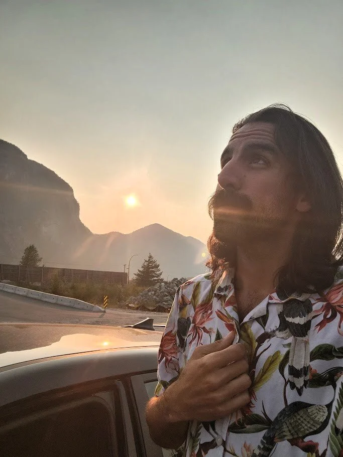 Daniel Deal  with long hair and a mustache wearing a floral shirt, standing outdoors in front of a car, with mountains and the sun setting or rising in the background.