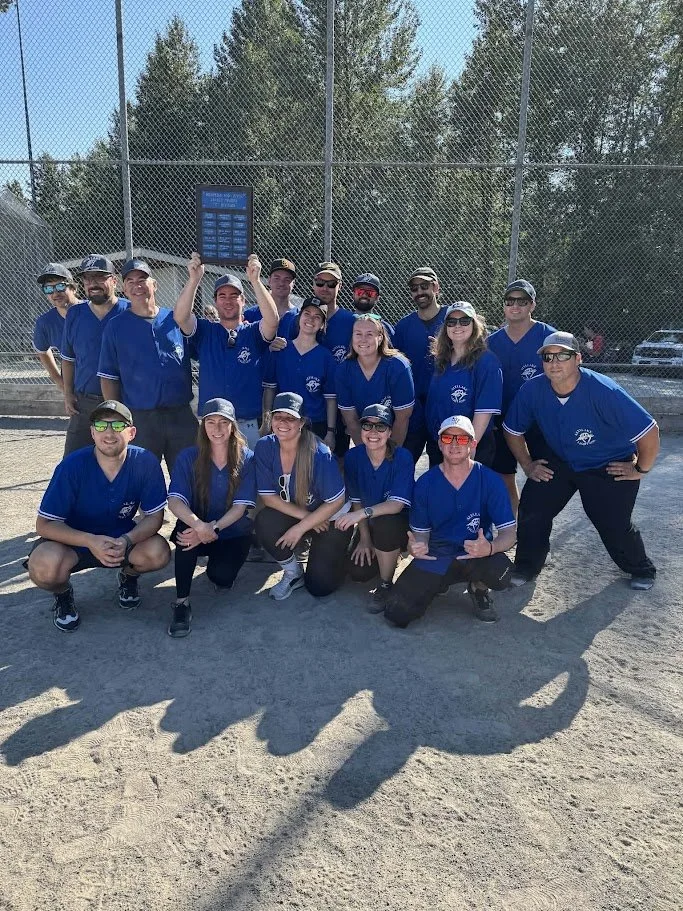 A group of people in matching blue baseball uniforms posing together on a baseball field, with a chain-link fence and trees in the background. Some are kneeling, others are standing, and one person in the back is holding a trophy.