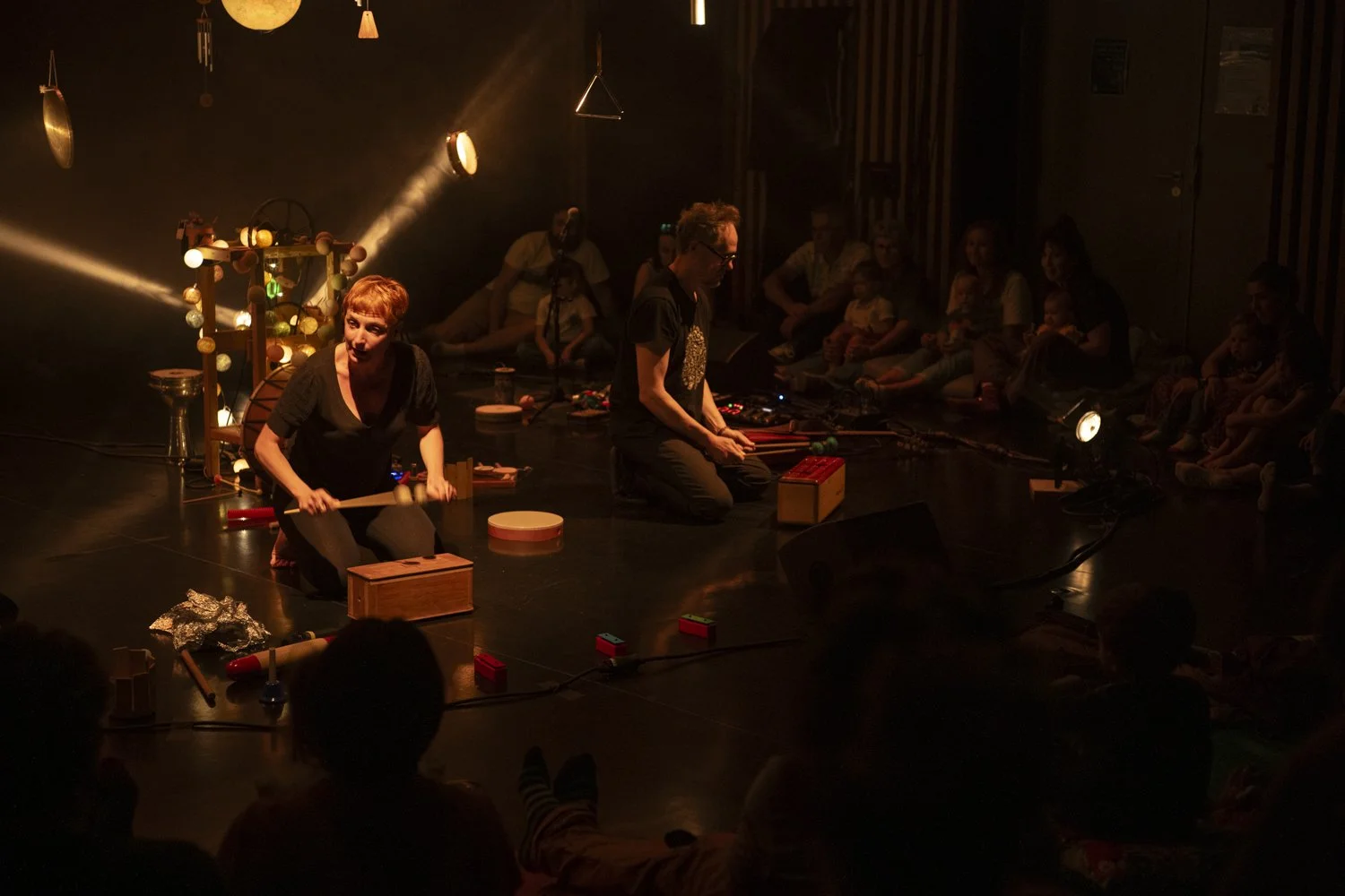 Une scène de spectacle avec des artistes jouant avec des instruments et des objets, entourés de spectateurs assis dans une ambiance tamisée, avec des éclairages dirigés vers la scène.