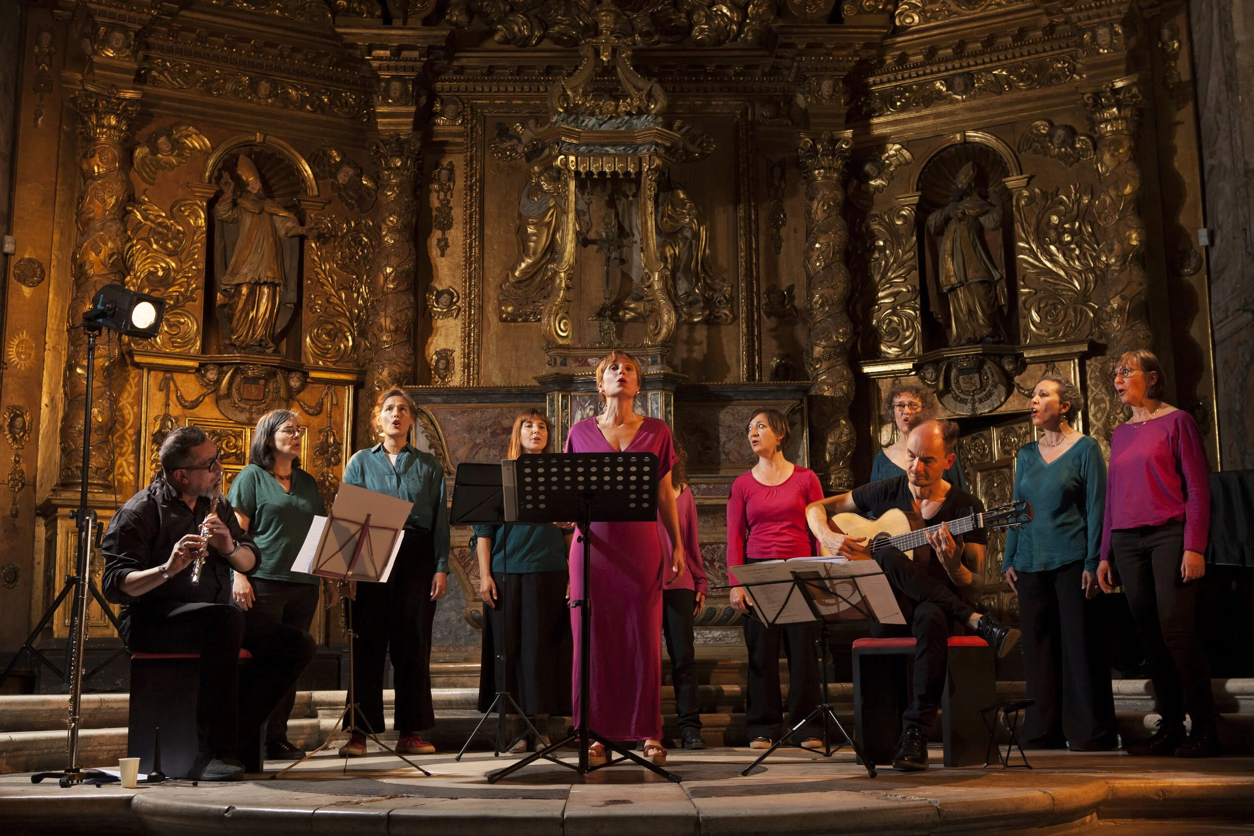 Un groupe de chanteurs et musiciens se produisent sur scène dans un théâtre ornée de sculptures dorées et de décorations baroques.