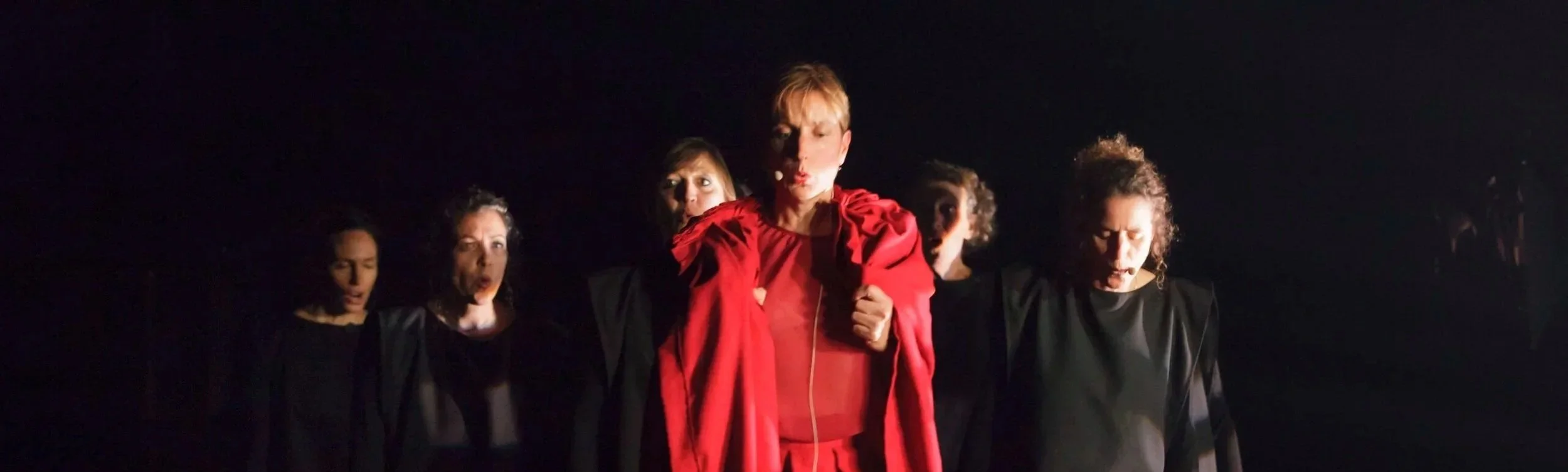 Groupe de six femmes en tenue sombre, avec la femme au centre en rouge, dans une scène sombre, probablement une performance artistique ou théâtrale.