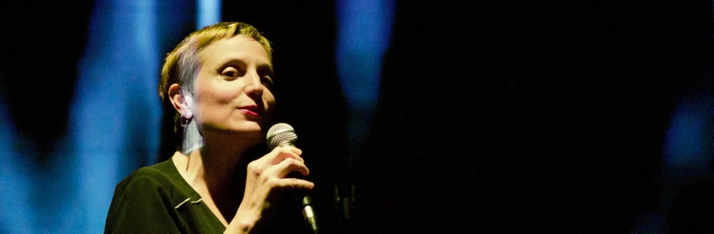 Une femme parle dans un microphone lors d'une performance ou d'une présentation, fond sombre avec lumière ciblant le visage.
