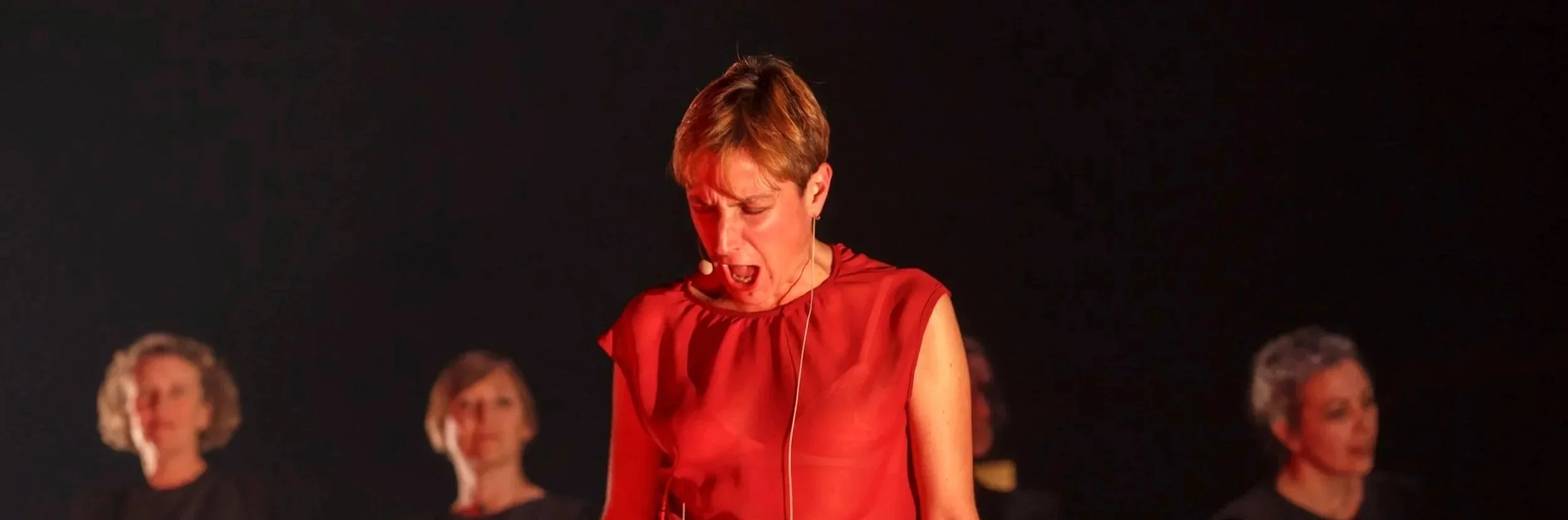 Une femme en rouge chante avec émotion lors d'une performance, entourée de femmes en noir dans un décor sombre.