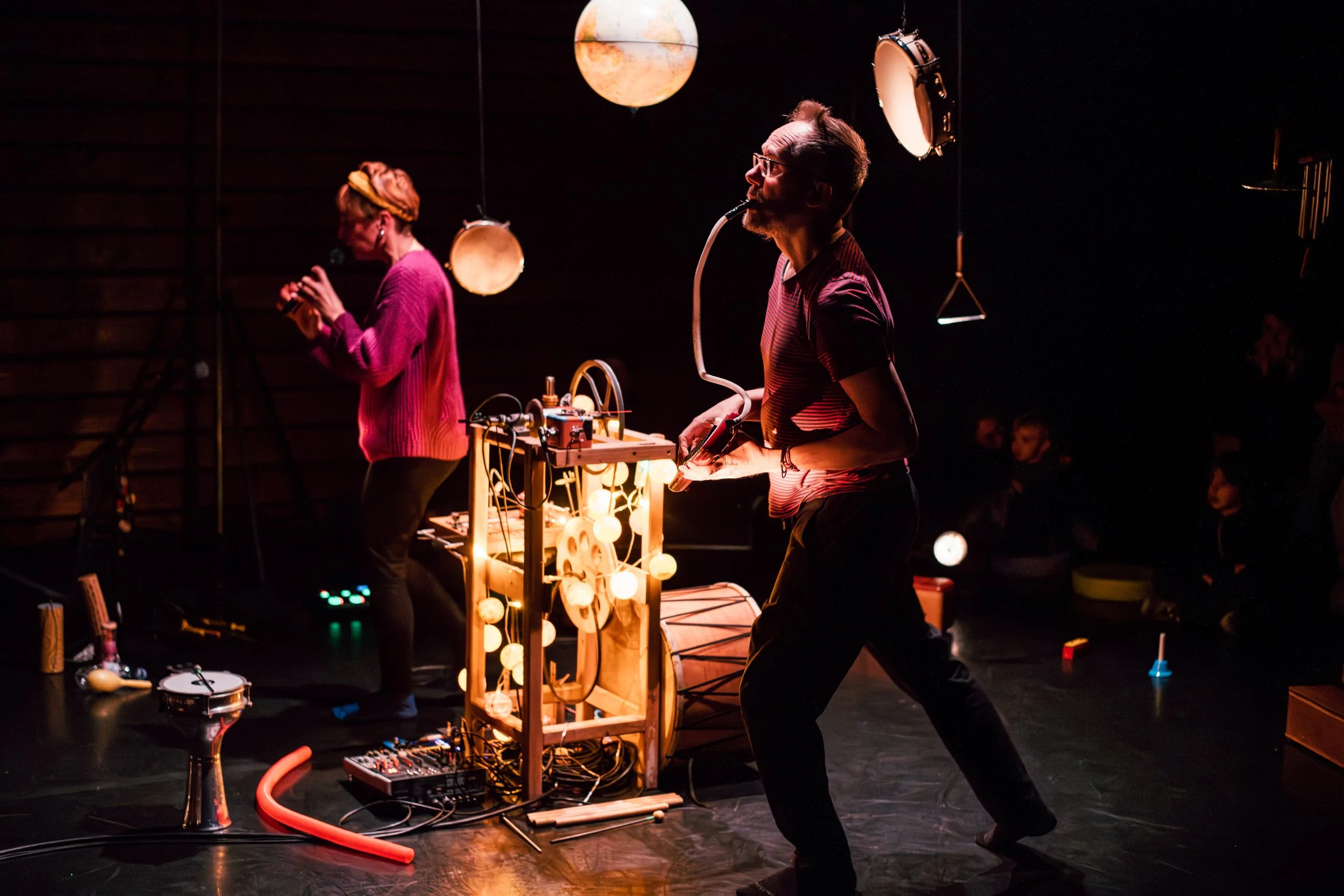 Deux personnes sur scène lors d'une performance artistique, avec un fond sombre, entourées d'équipements de musique et de lumière, dont des lampes lumineuses suspendues et un globe lumineux.