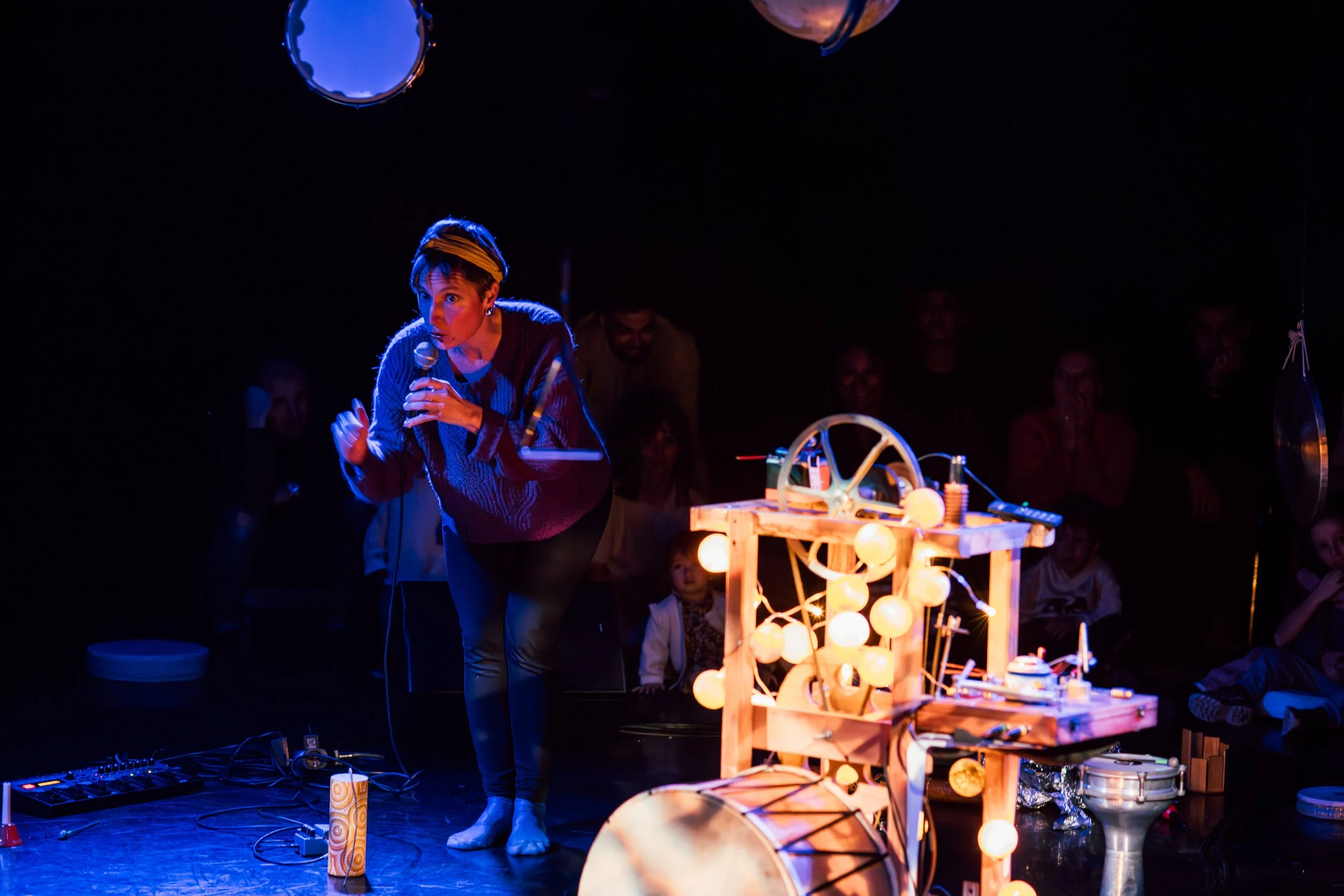 Une femme avec un bandeau sur la tête en train de parler ou de chanter lors d'une performance artistique ou théâtrale, entourée d'un public assis dans l'obscurité, avec une installation lumineuse en bois et boules lumineuses