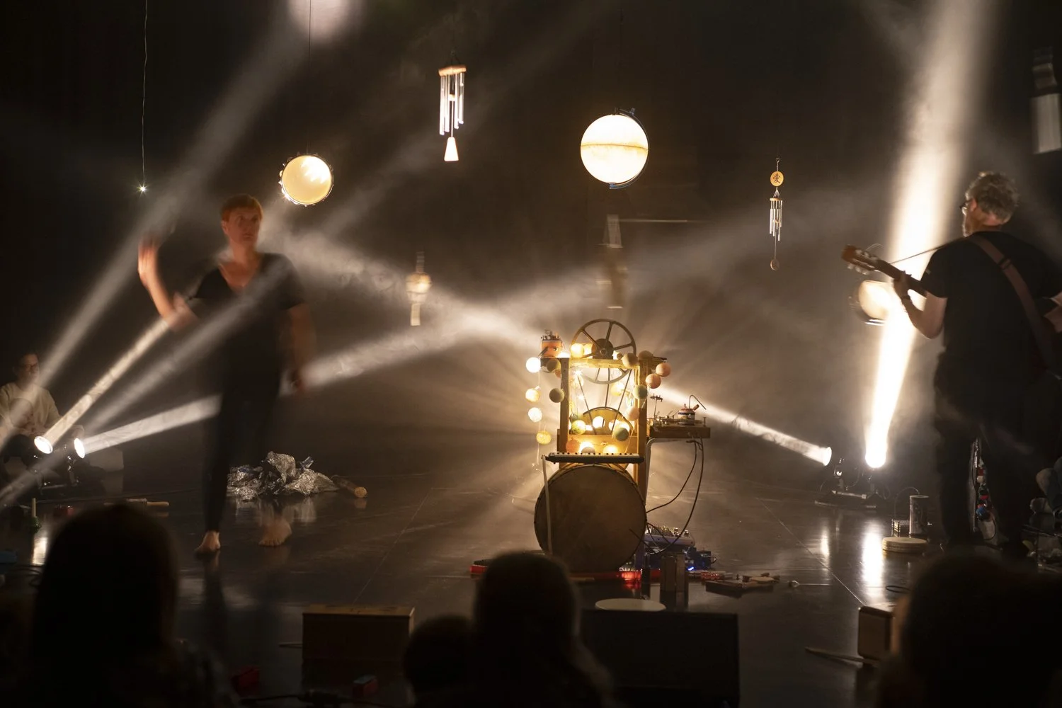 Scène artistique avec deux musiciens, un homme jouant de la guitare et une femme dansant, entourés de lumières et d'objets suspendus au plafond lors d'une performance.