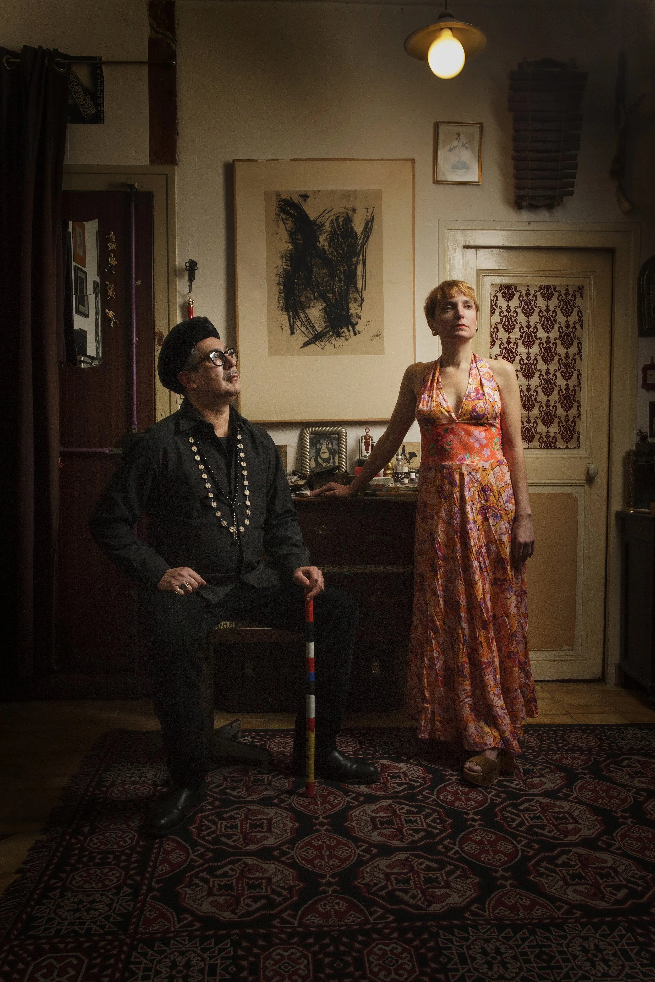 Deux personnes posent dans un intérieur avec des œuvres d'art sur les murs. La personne à gauche porte une chemise noire, des lunettes, une chaîne de perles, un béret noir, et tient un bâton coloré. La personne à droite porte une robe noire, une robe