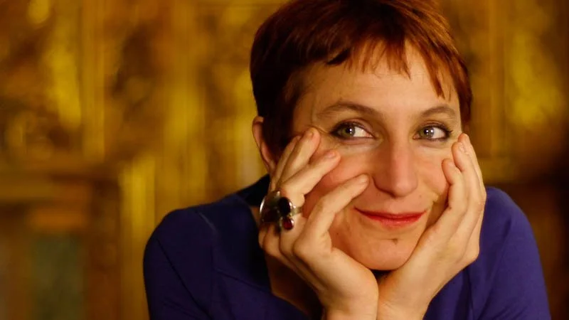 Une femme avec des cheveux courts et roux, portant un blazer violet, sourit en posant ses mains sur son visage, dans un décor chaud et flou.