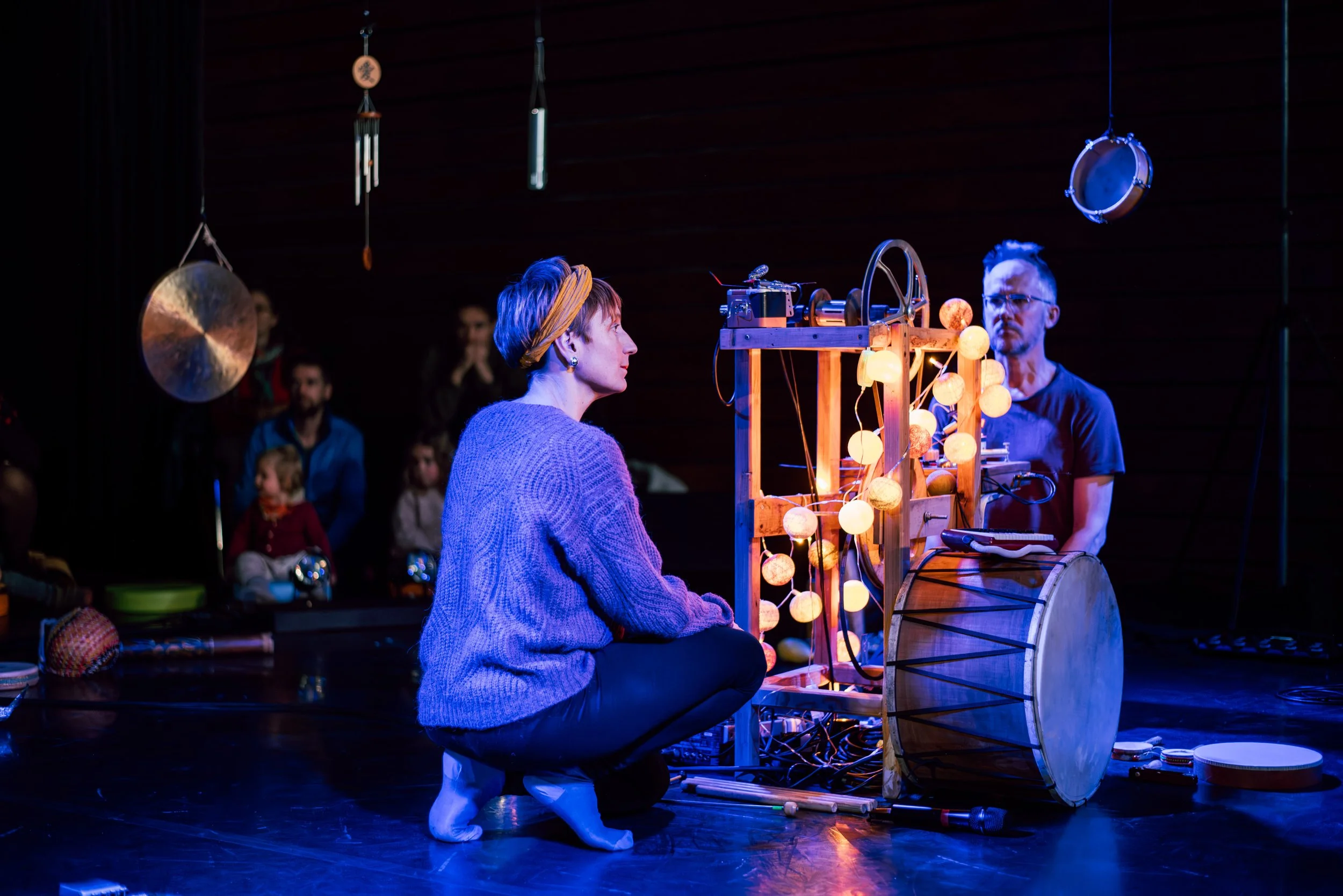 Une femme au sommet, une personne assise, observe un dispositif lumineux en forme d'arc et d'ampoules, lors d'une performance ou atelier artistique.