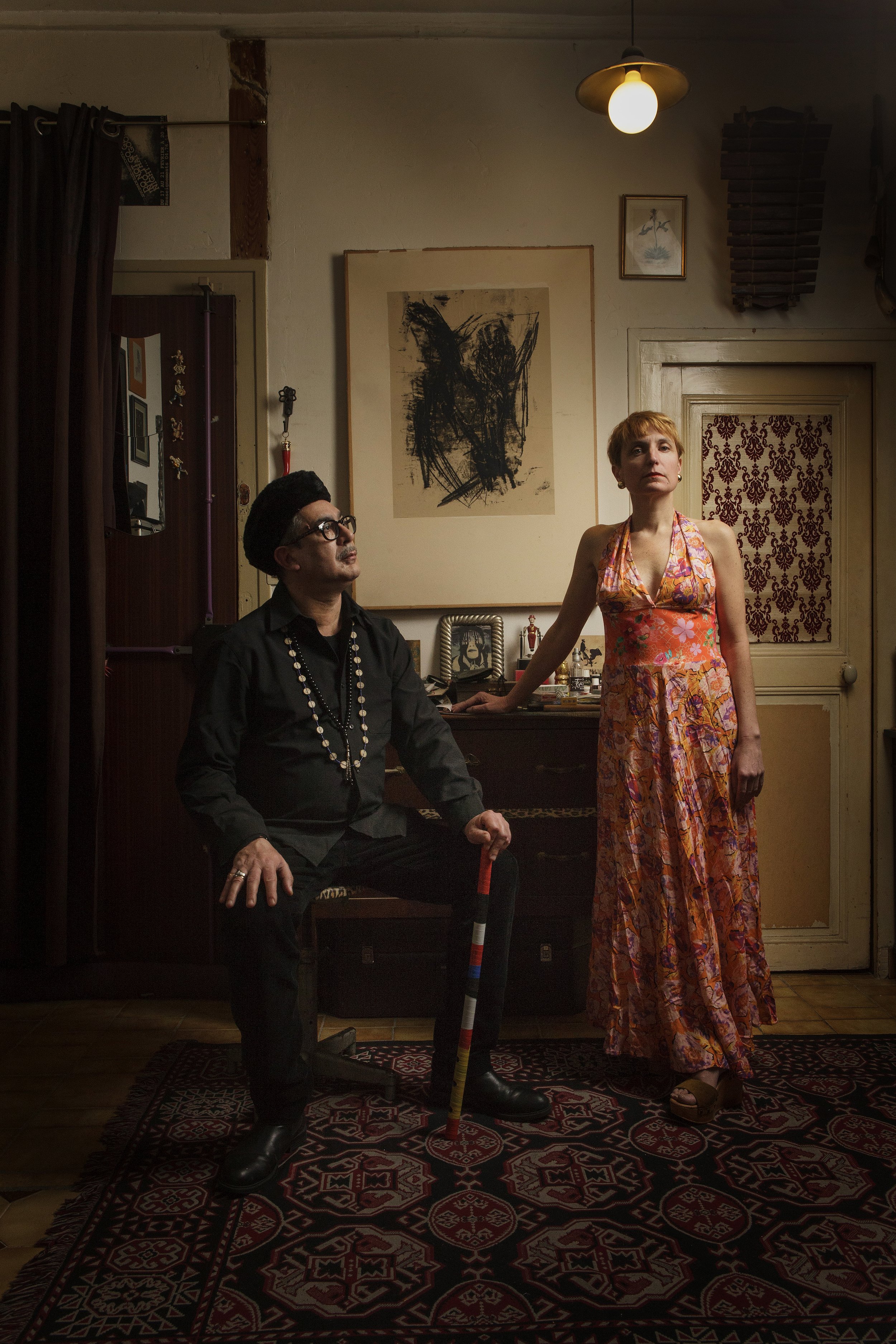 Deux personnes dans une pièce décorée avec des œuvres d'art. L'une est assise, portant des lunettes, un chapeau noir et une tenue noire avec un collier de perles, l'autre est debout, portant une robe longue à motifs floraux orange et rose. La pièce a