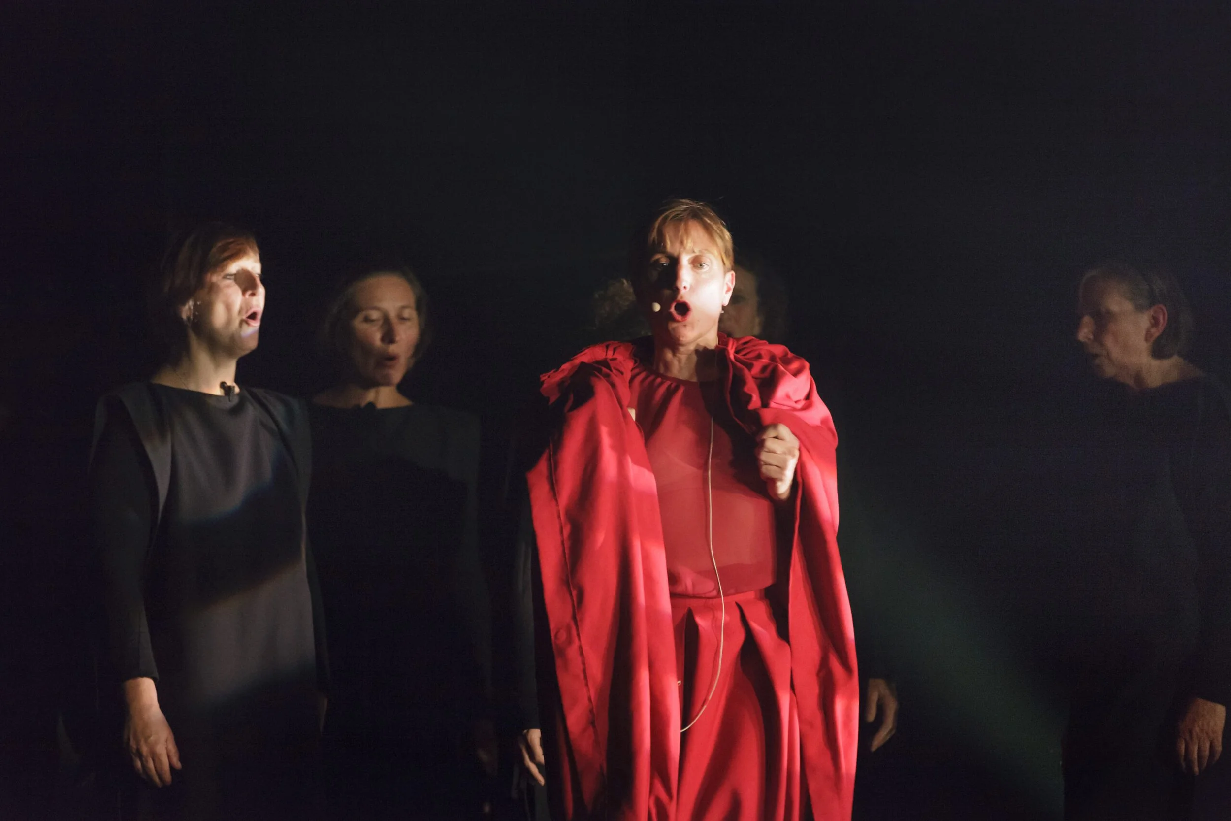 Groupe de personnes chantant ou récitant avec émotions, éclairage sombre, personne au centre en robe rouge. 