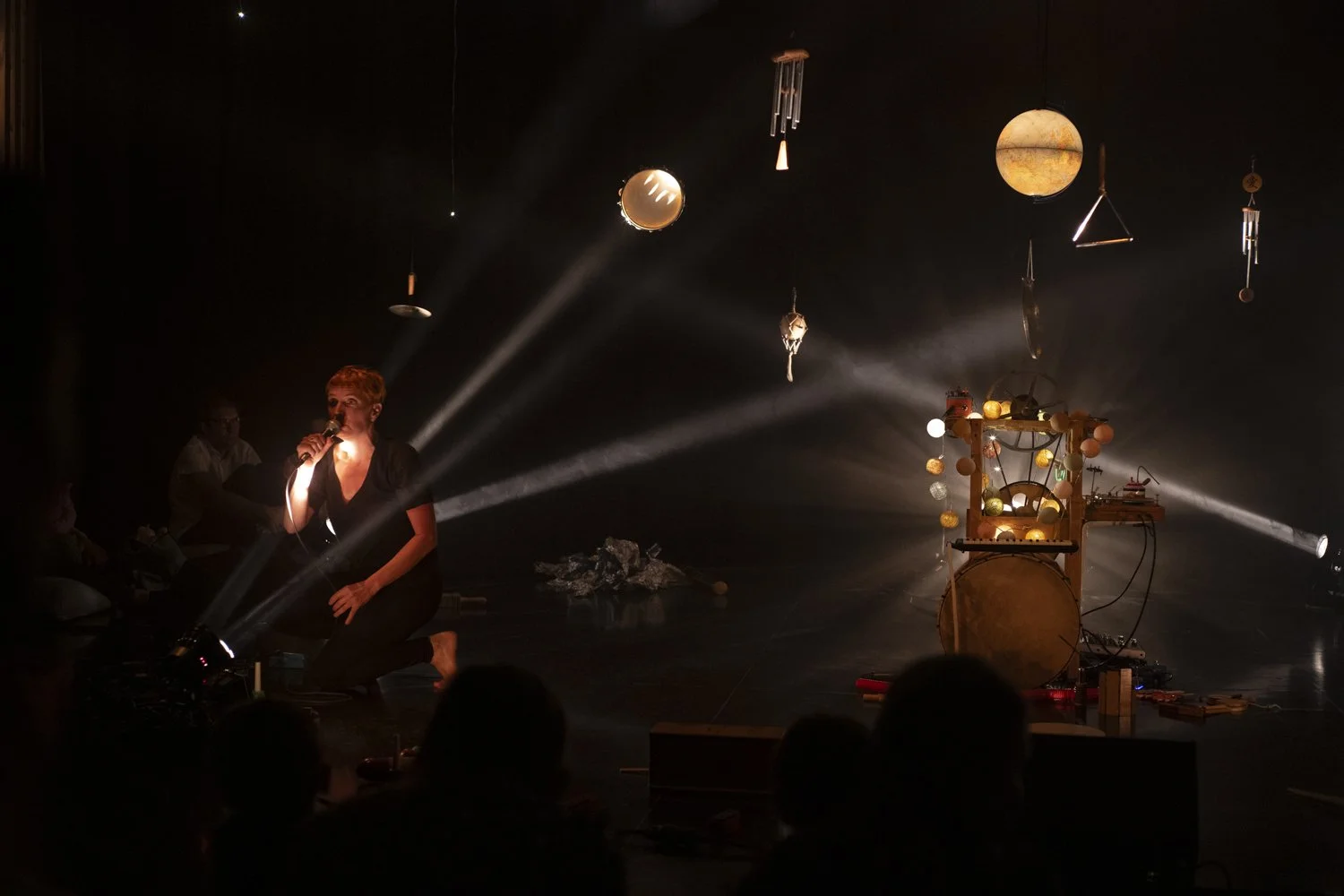 Une femme chante avec un microphone sur scène, entourée d'effets lumineux et de décorations suspendues, dont des sphères et un instrument mystérieux avec des lumières, dans une atmosphère sombre et artistique.
