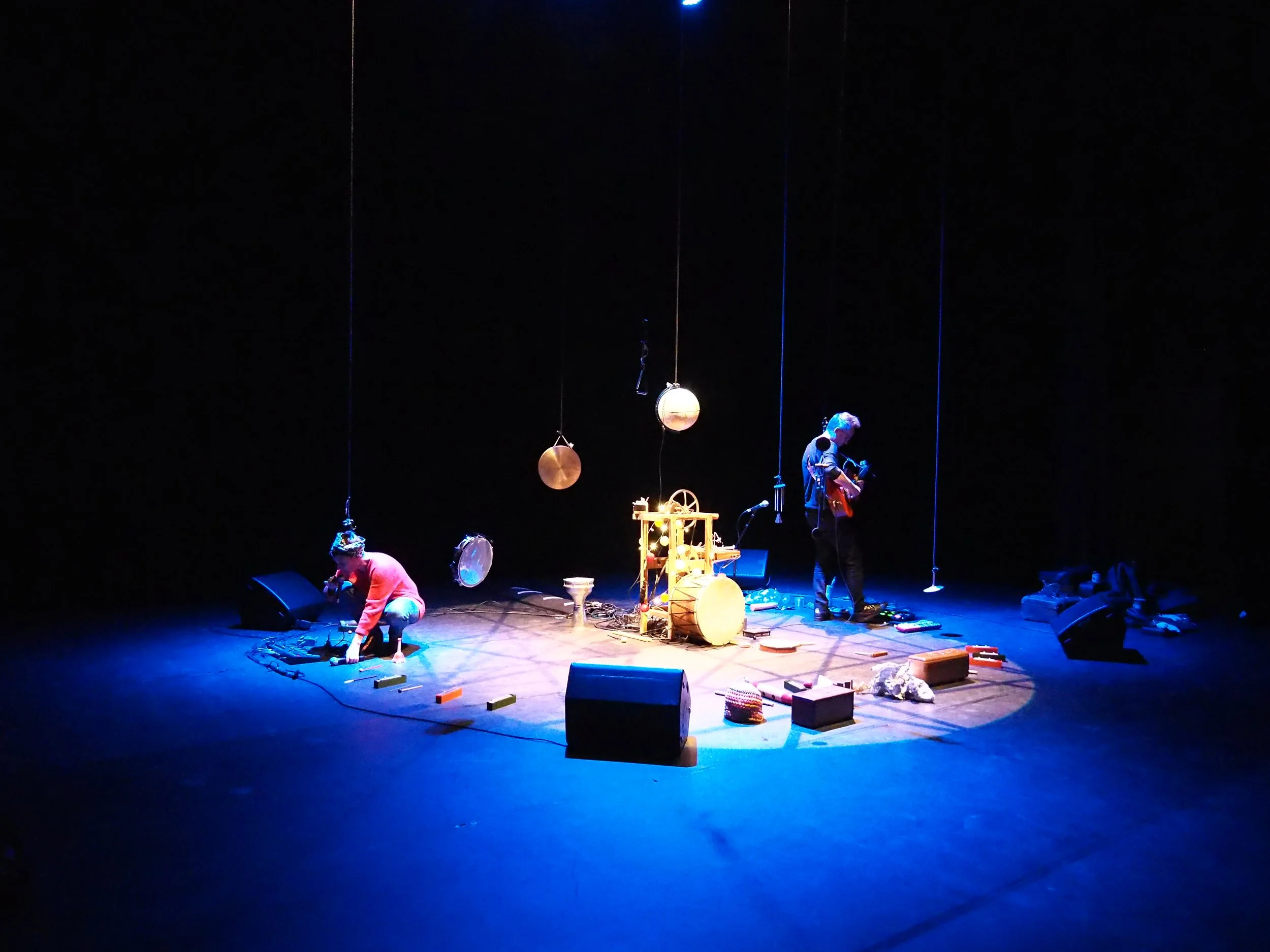 Une scène de spectacle avec deux artistes sur scène, un jouant de la guitare, entourés d'instruments de musique et d'accessoires dispersés, sous un éclairage tamisé.