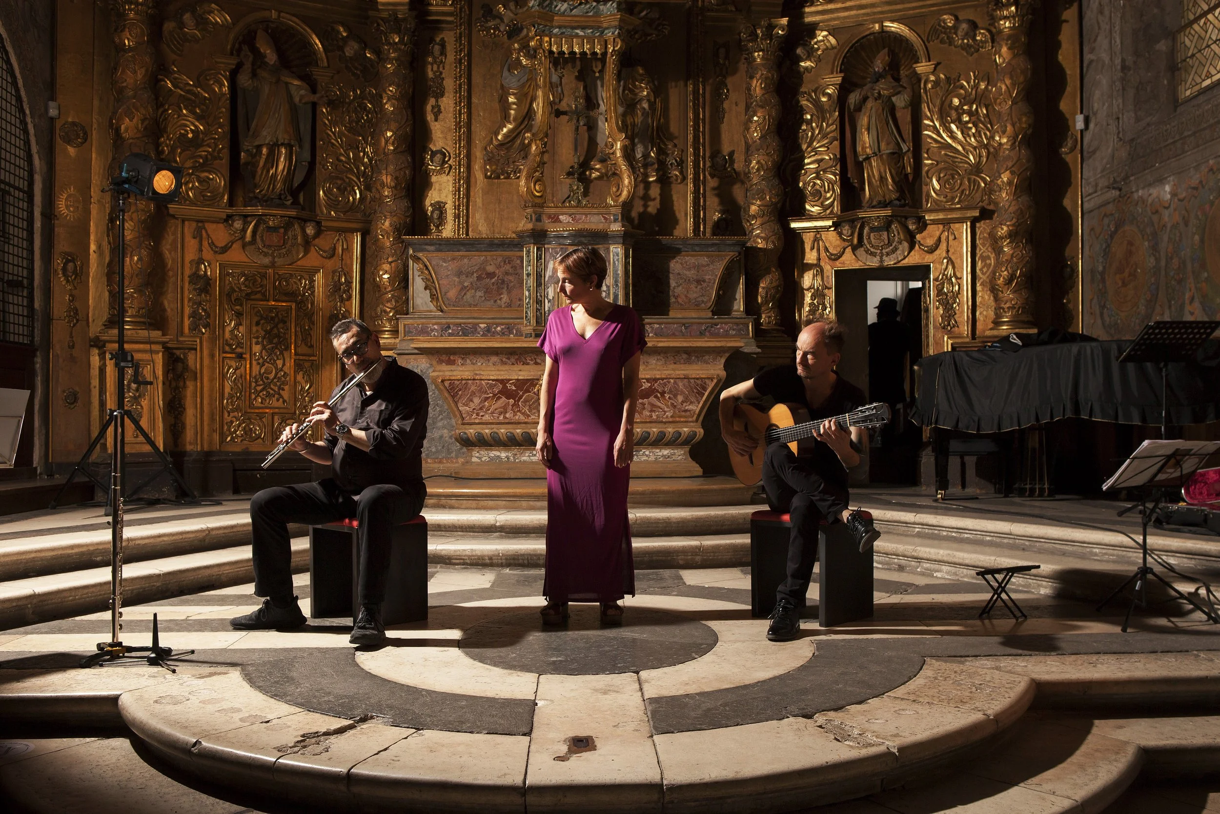Trois musiciens et une chanteuse en performance dans une salle ornée de dorures et de sculptures, avec un piano et des micros.